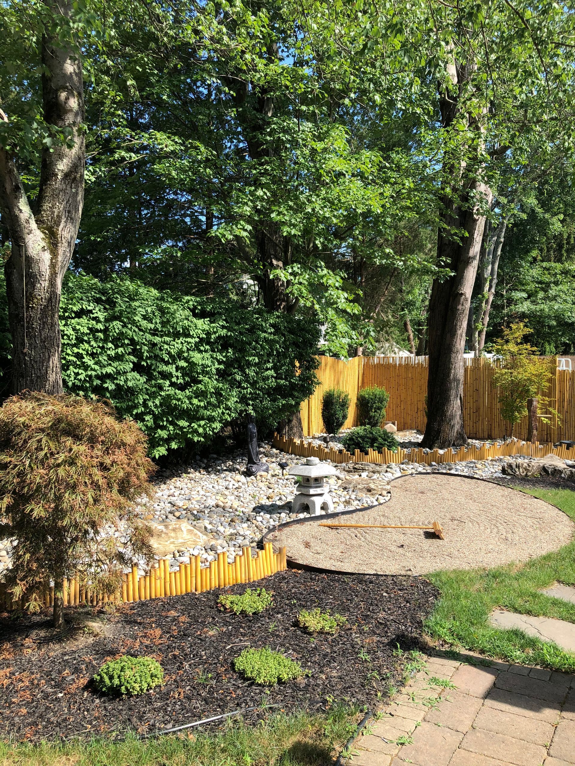 Backyard garden with trees, mulch beds, stones, and a small gravel path bordered by a wooden fence