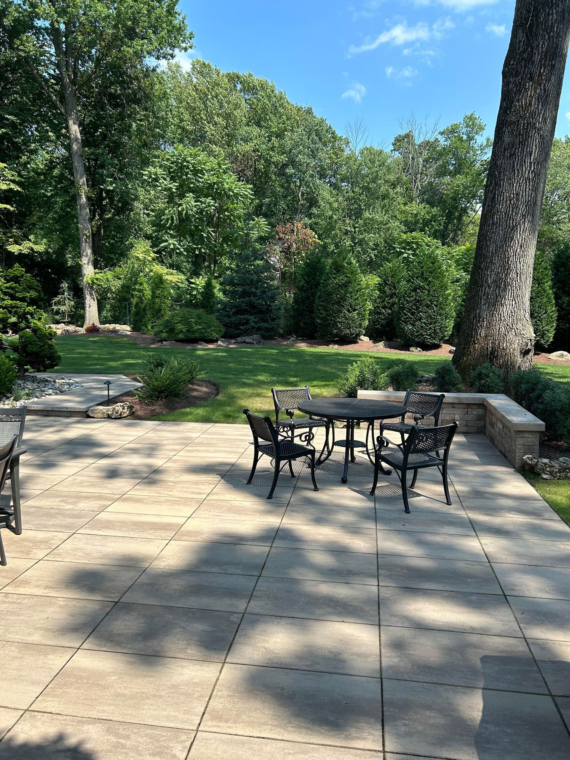 Patio with black metal table and chairs beside a green garden and stone steps in sunlight