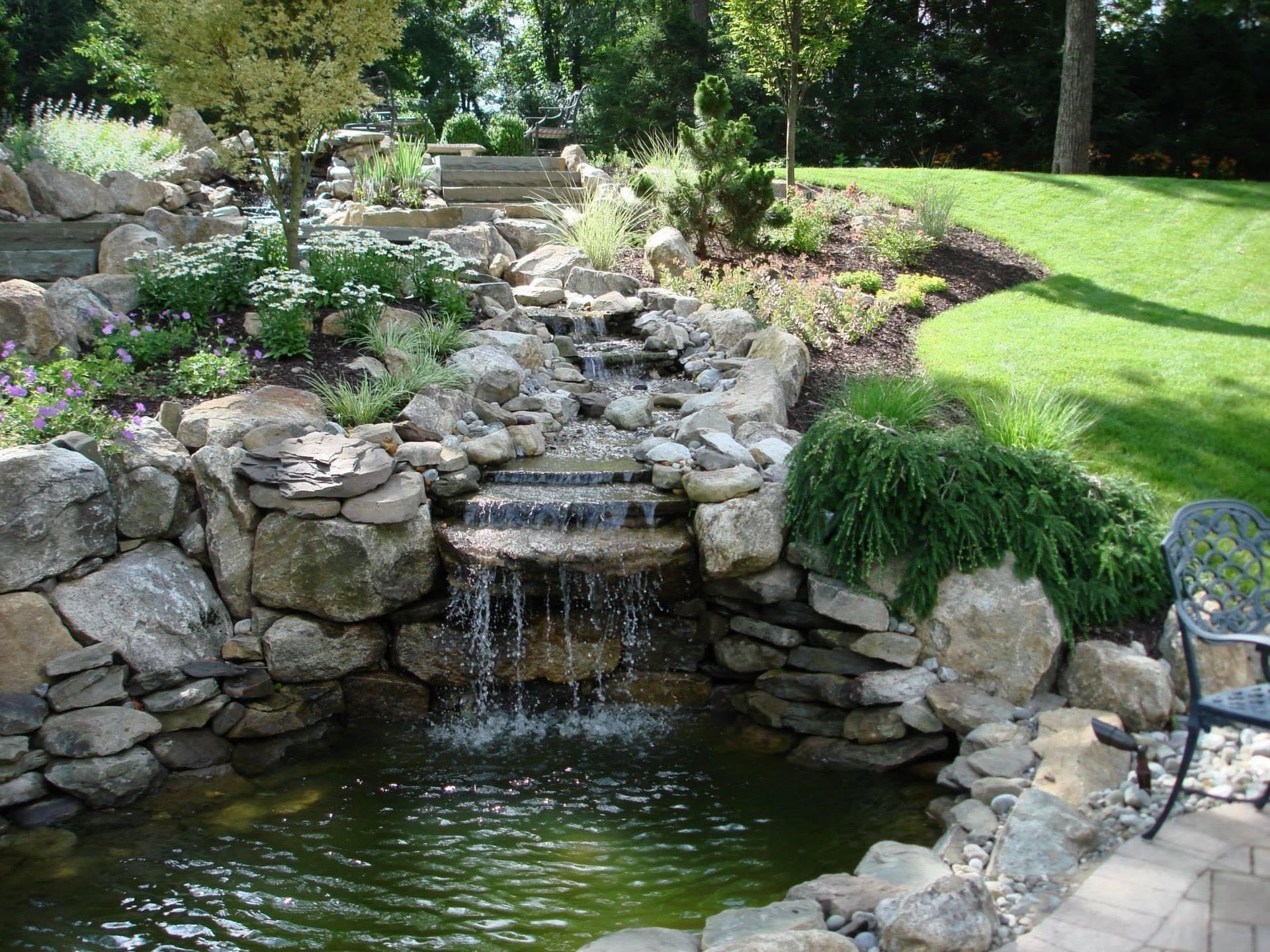 Stone backyard waterfall flowing into a small pond beside a grassy lawn and patio chair
