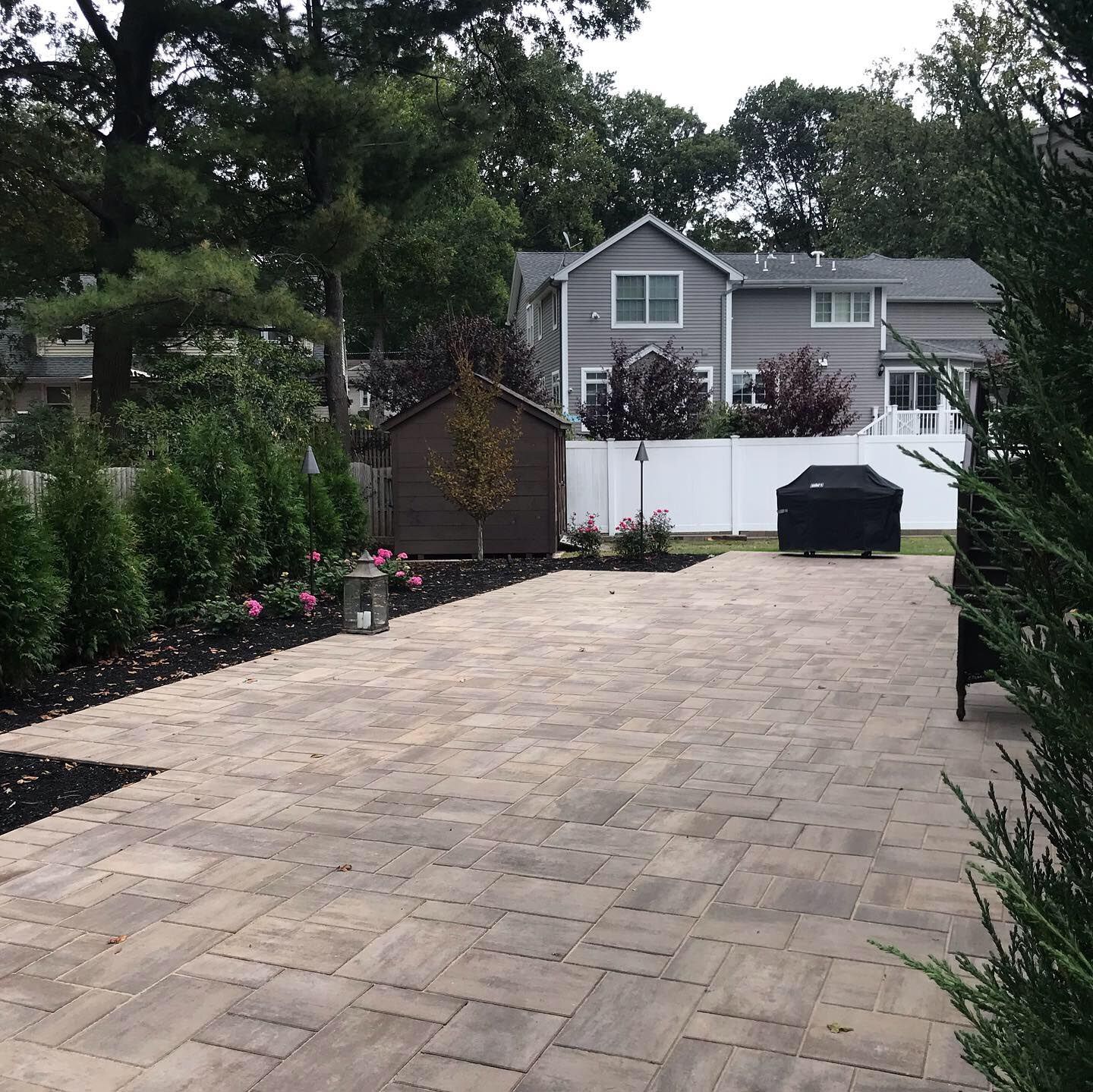 Stone-paved driveway leading to a gray house, bordered by shrubs and a white fence, with a black bin at the end.