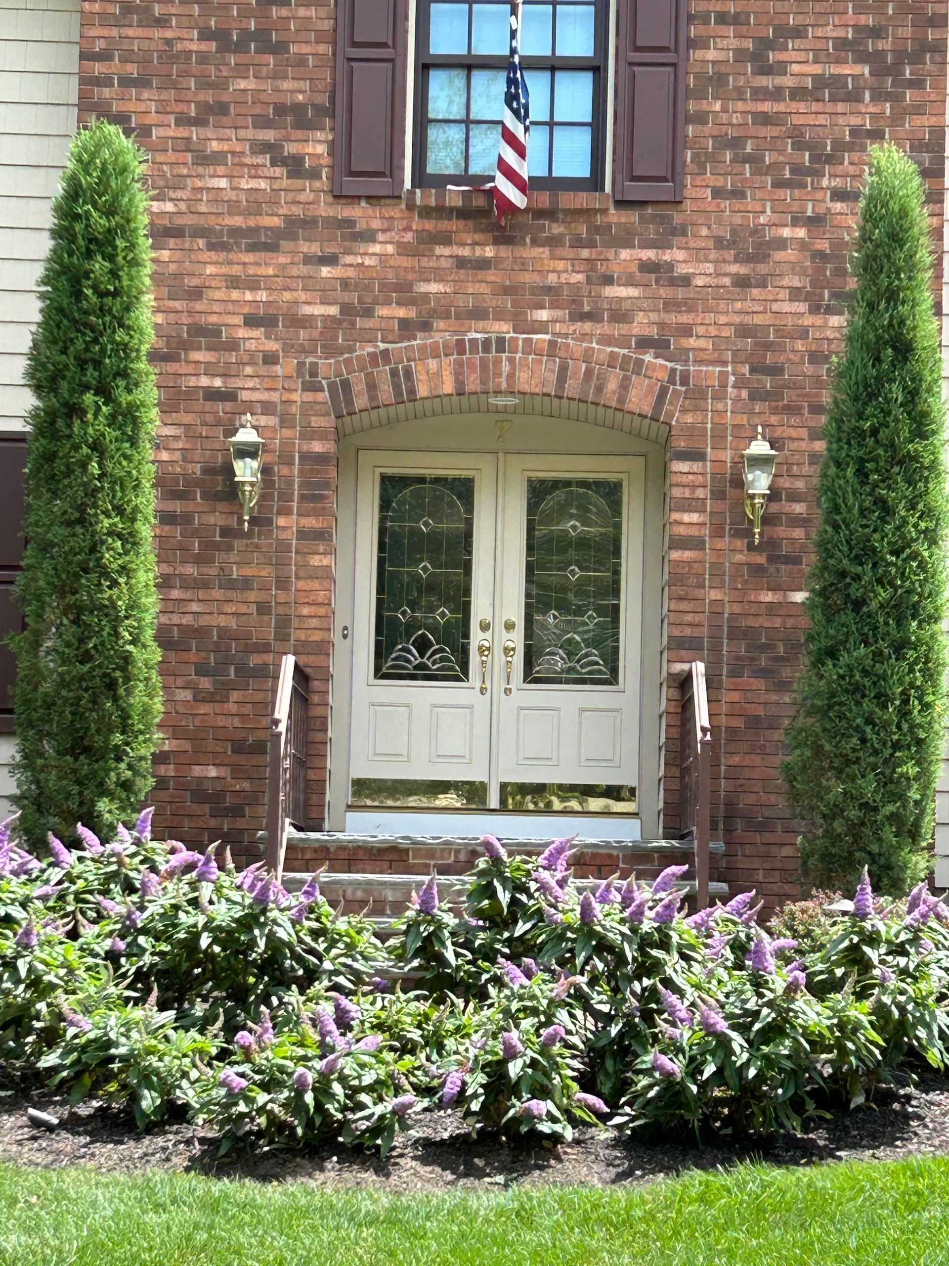 Brick house entrance with white double doors, tall shrubs, and purple flowers in front yard