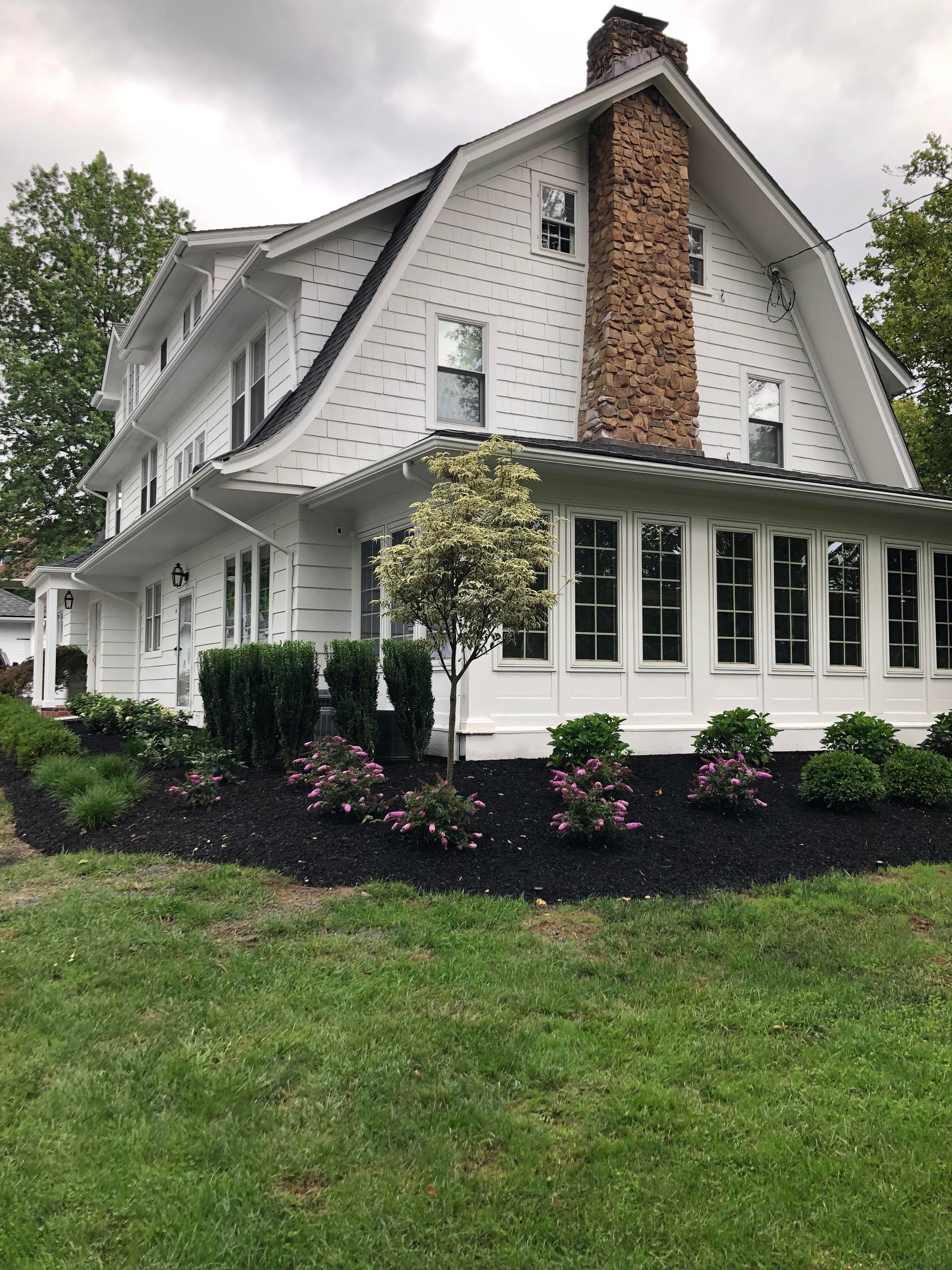 White farmhouse with black shutters and brick chimney, surrounded by manicured lawn and shrubs.
