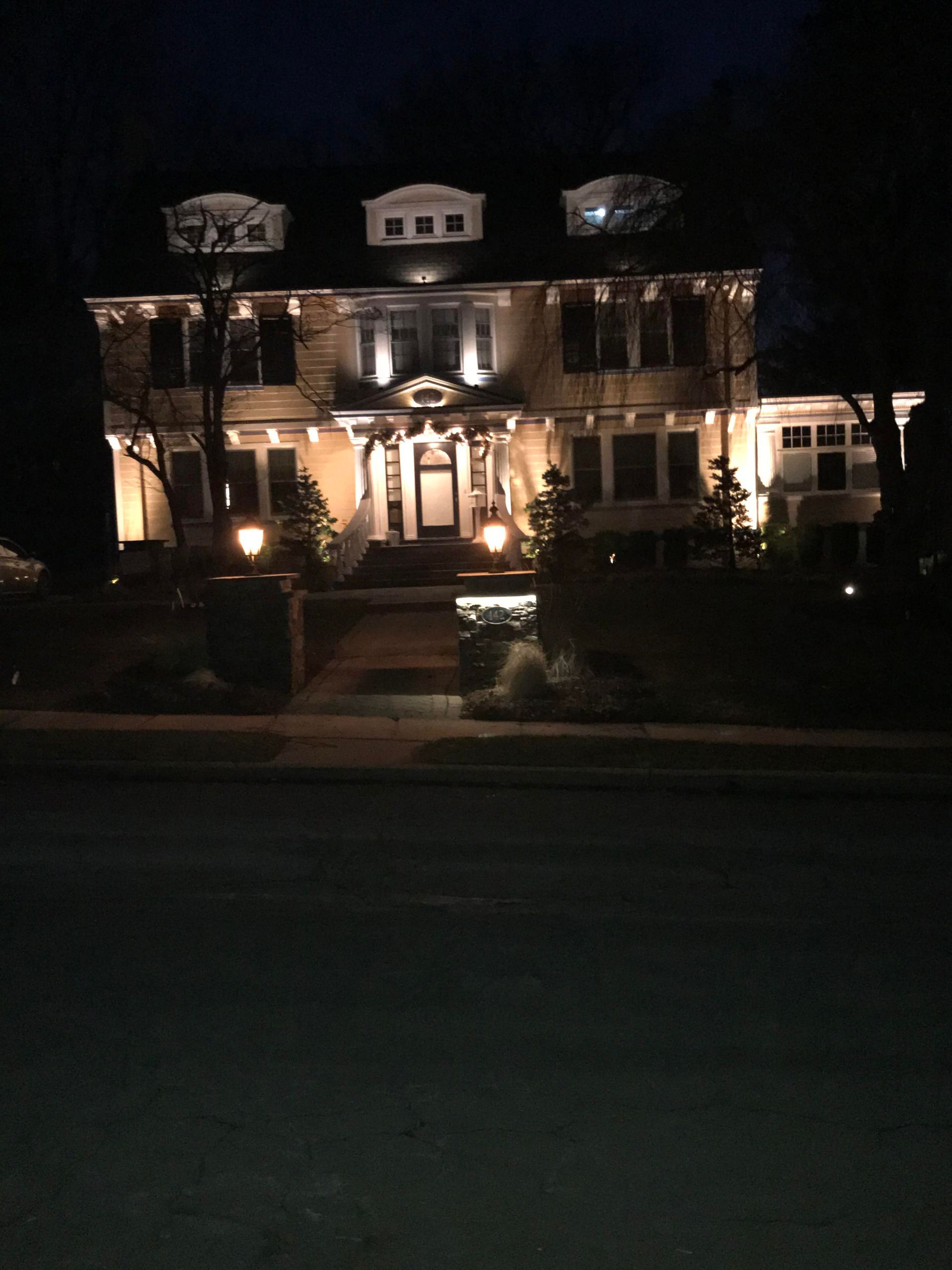Nighttime view of a warmly lit two-story house with front porch, steps, and dark lawn at the street.