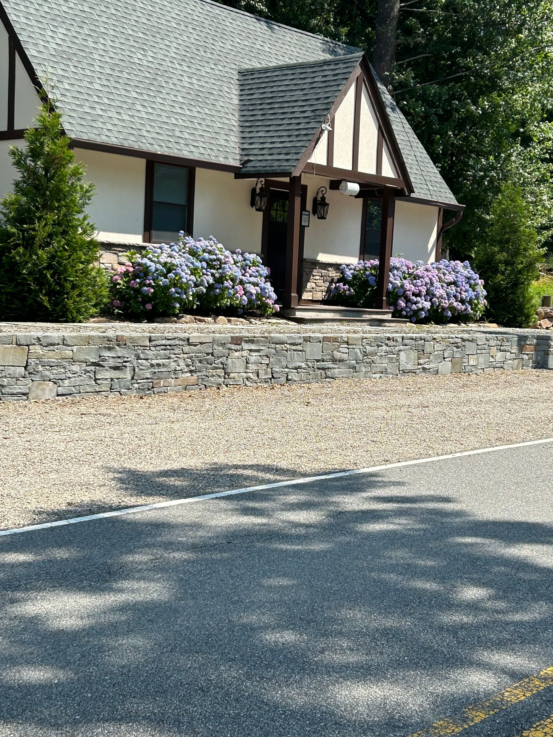 Stone house with a steep shingled roof, flower beds, and a gravel driveway in bright sunlight