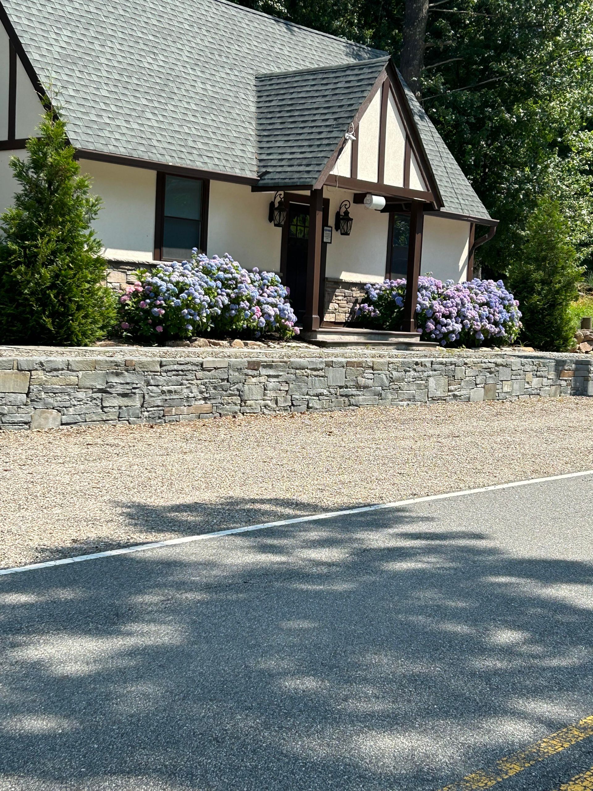 Cottage with steep shingled roof, stone retaining wall, and blooming shrubs beside a gravel drive.