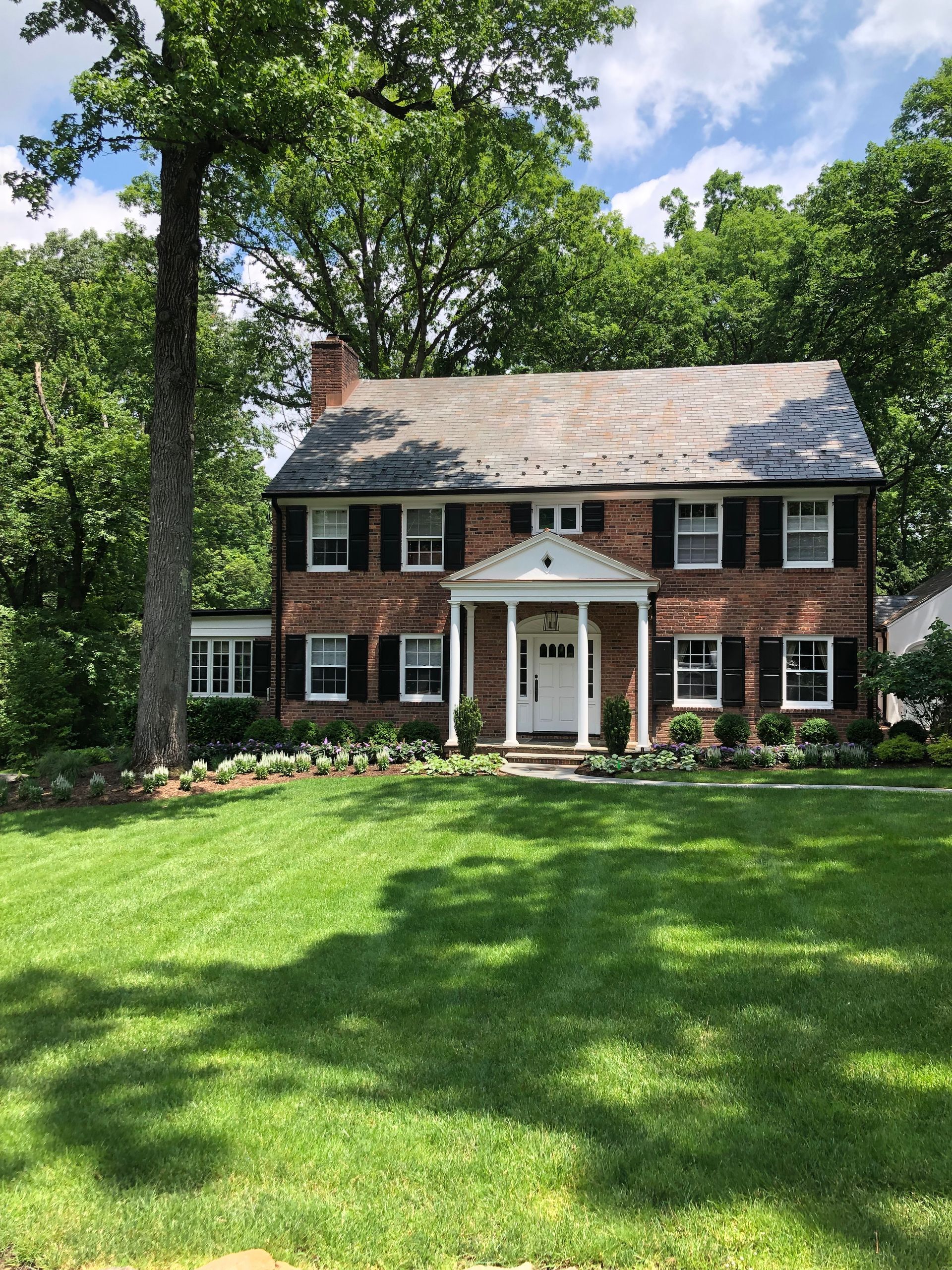 Brick colonial house with white trim and large green lawn under sunny trees