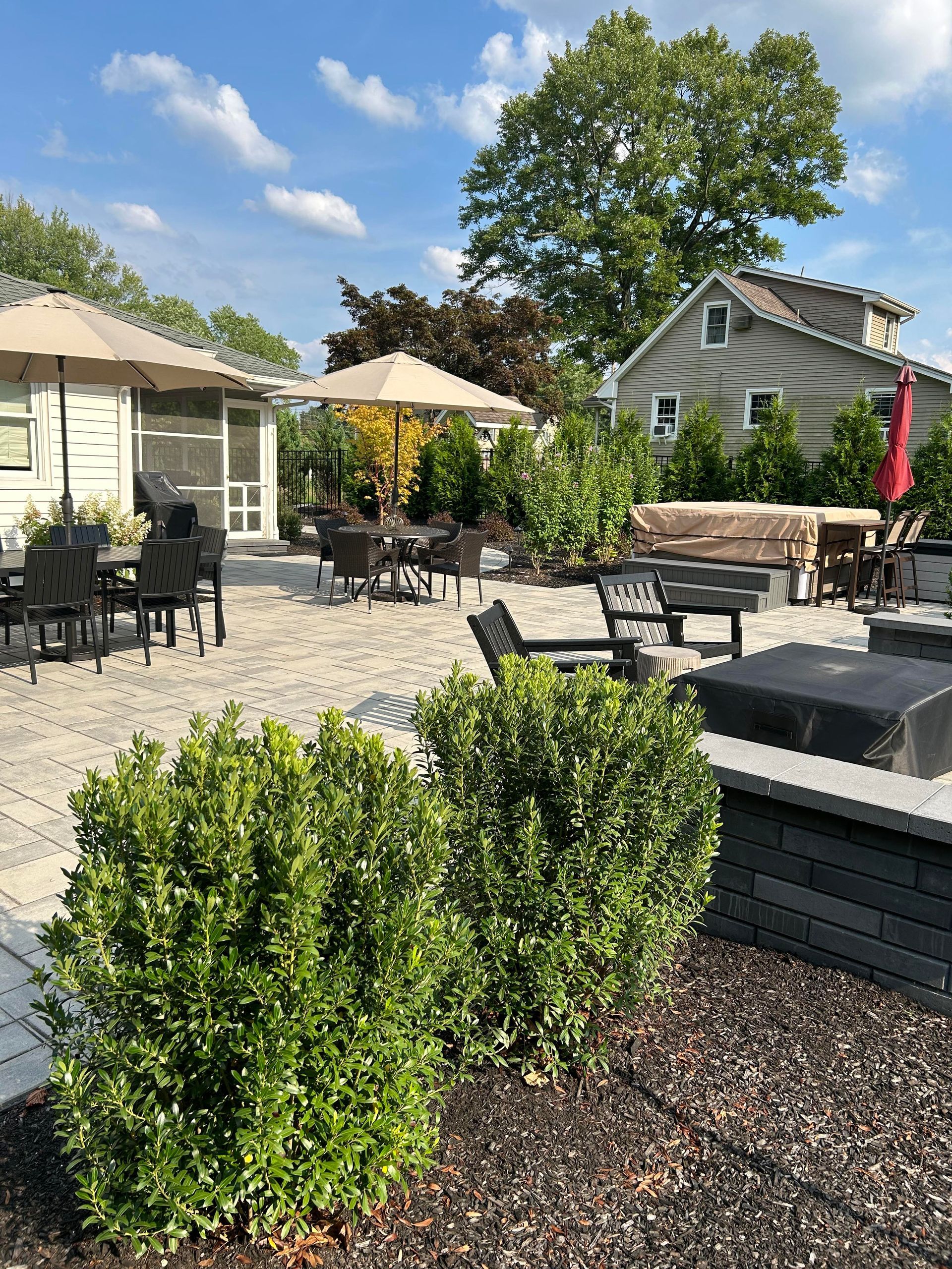 Patio with black chairs, tan umbrellas, potted shrubs, and stone planters in a sunny garden area