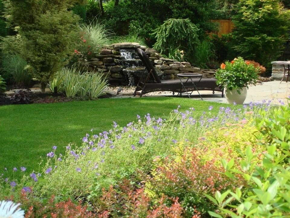 Lush garden with green lawn, purple flowers, patio chairs, and a small waterfall amid dense trees