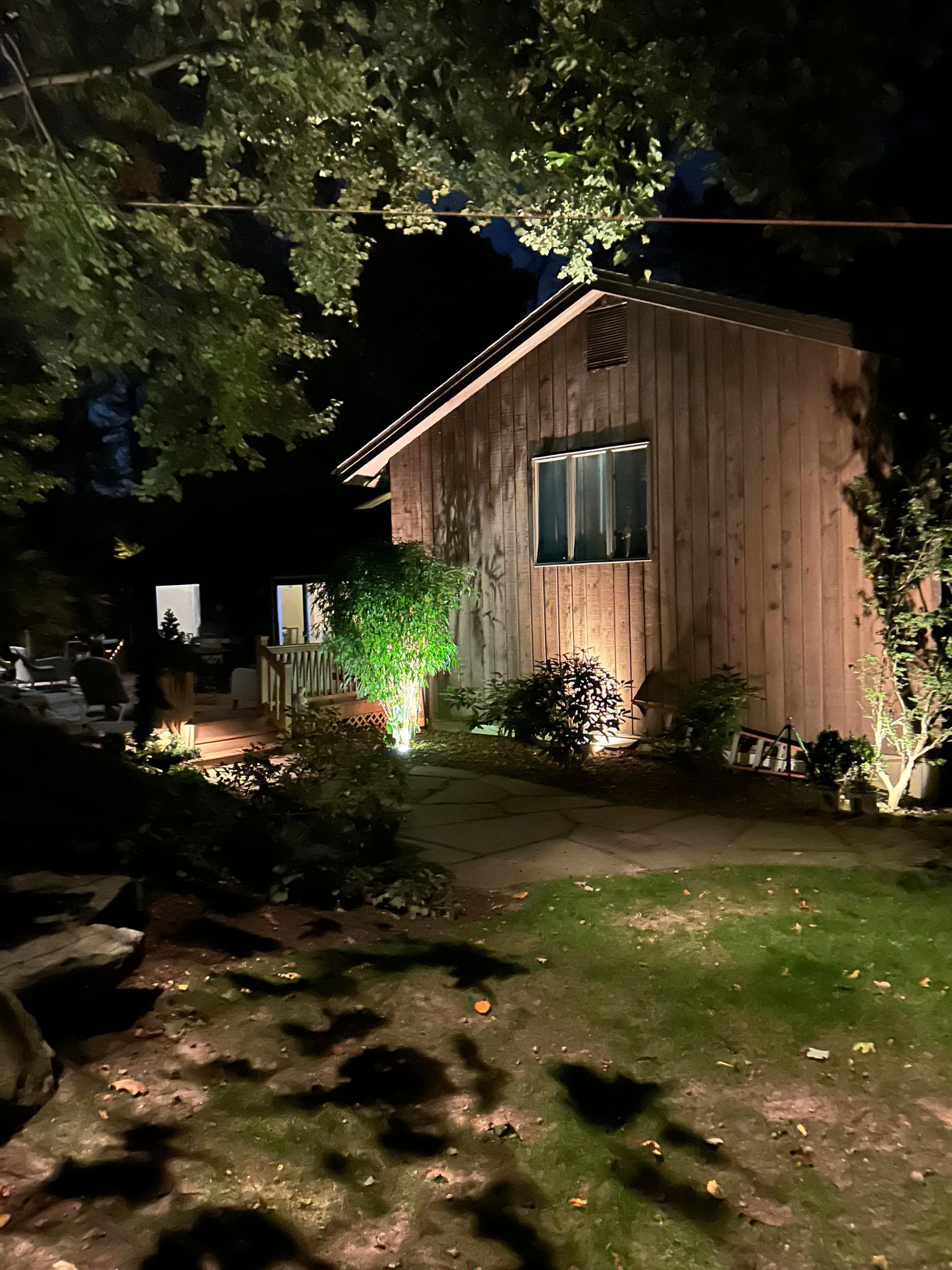 Nighttime garden view of a wooden cabin lit by outdoor lights, with trees, shrubs, and a grassy yard.