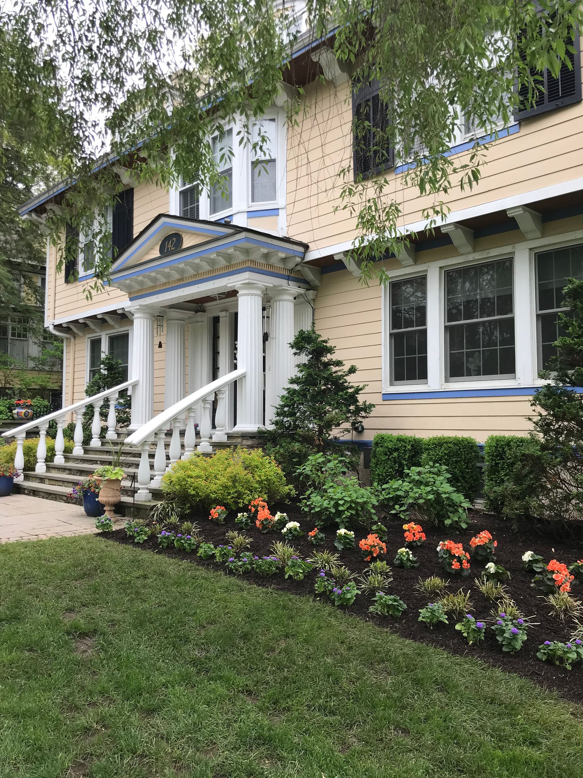 Two-story beige house with white porch and railings, landscaped front yard, and colorful flower beds.