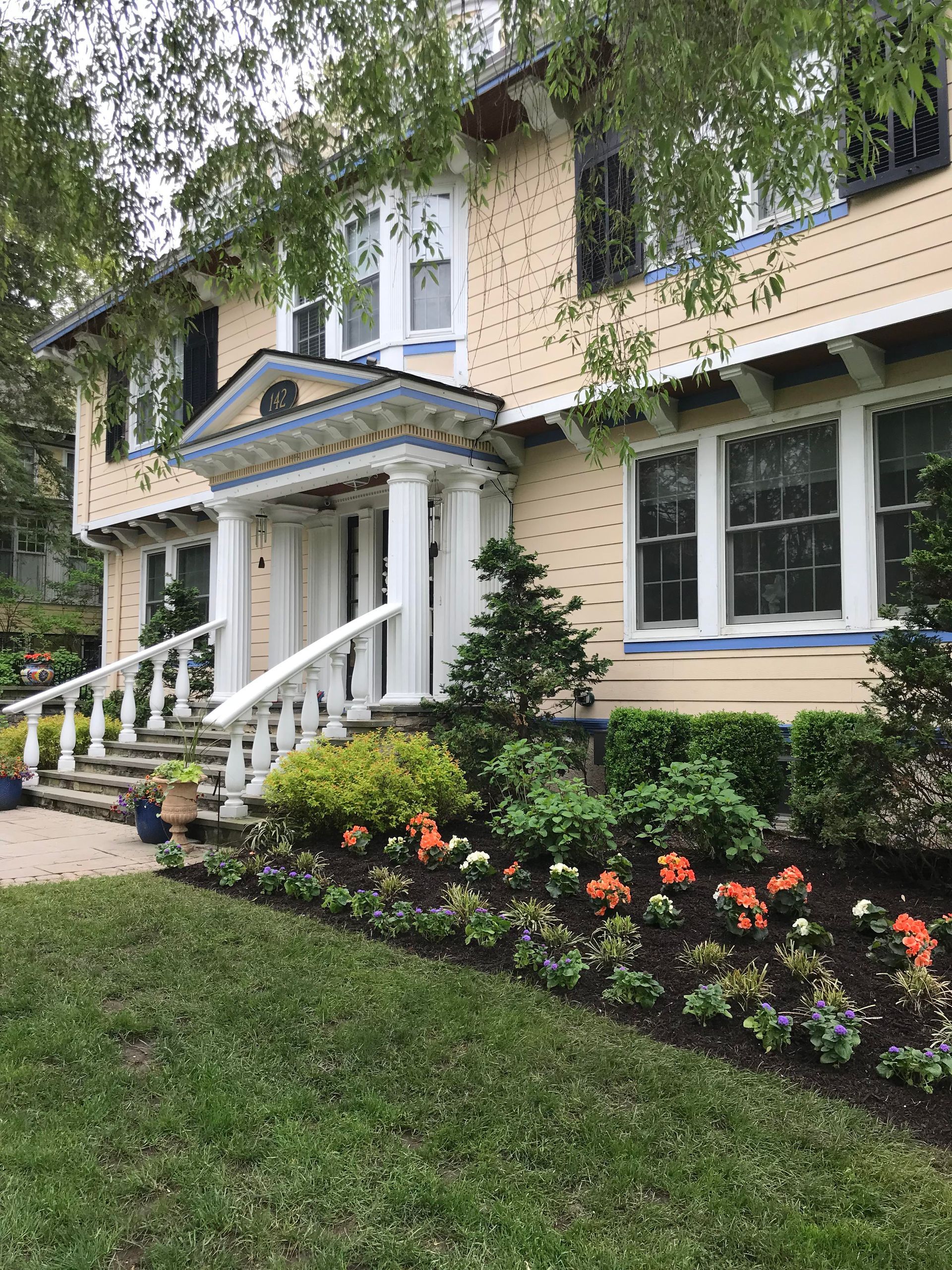 Yellow house with white porch, black-trimmed windows, and flower beds under hanging tree branches