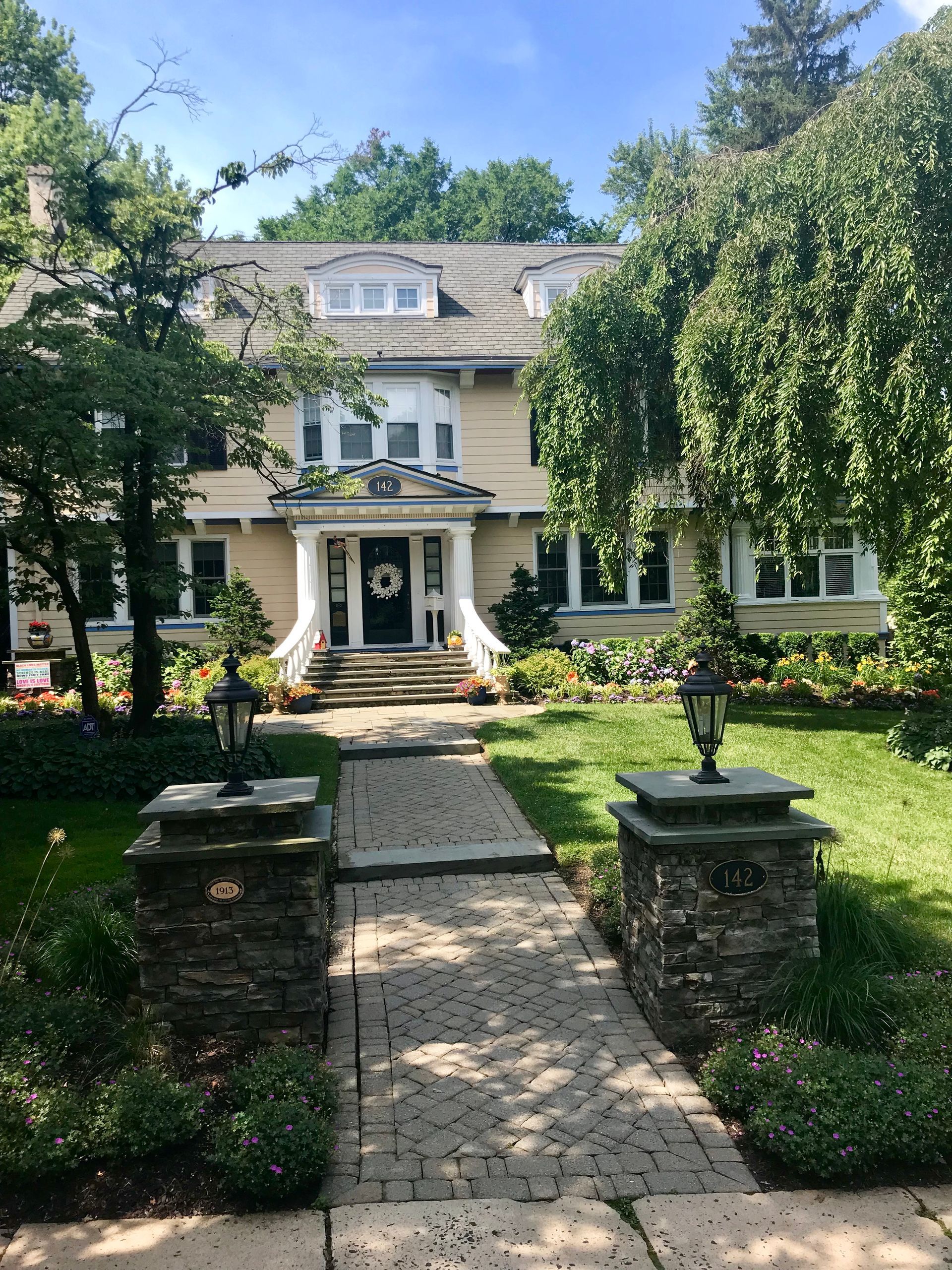 White two-story house with a front porch, stone walkway, and lush green trees and shrubs.