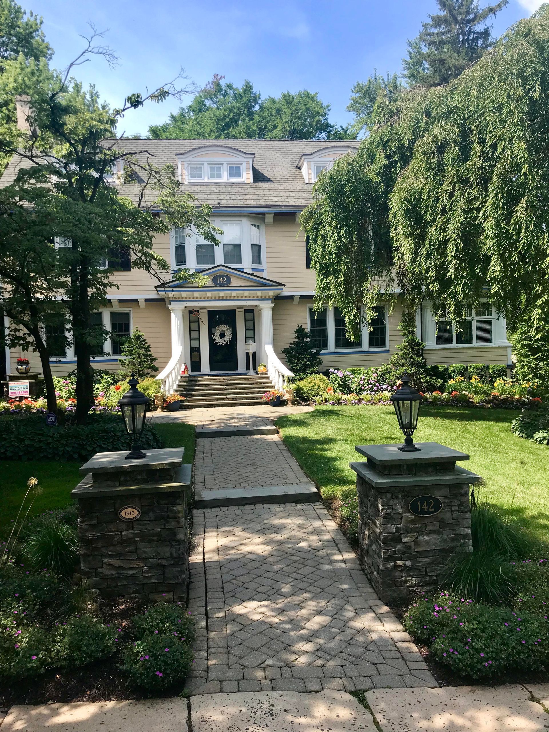 Large white house with a front walkway, garden, and trees on a sunny day