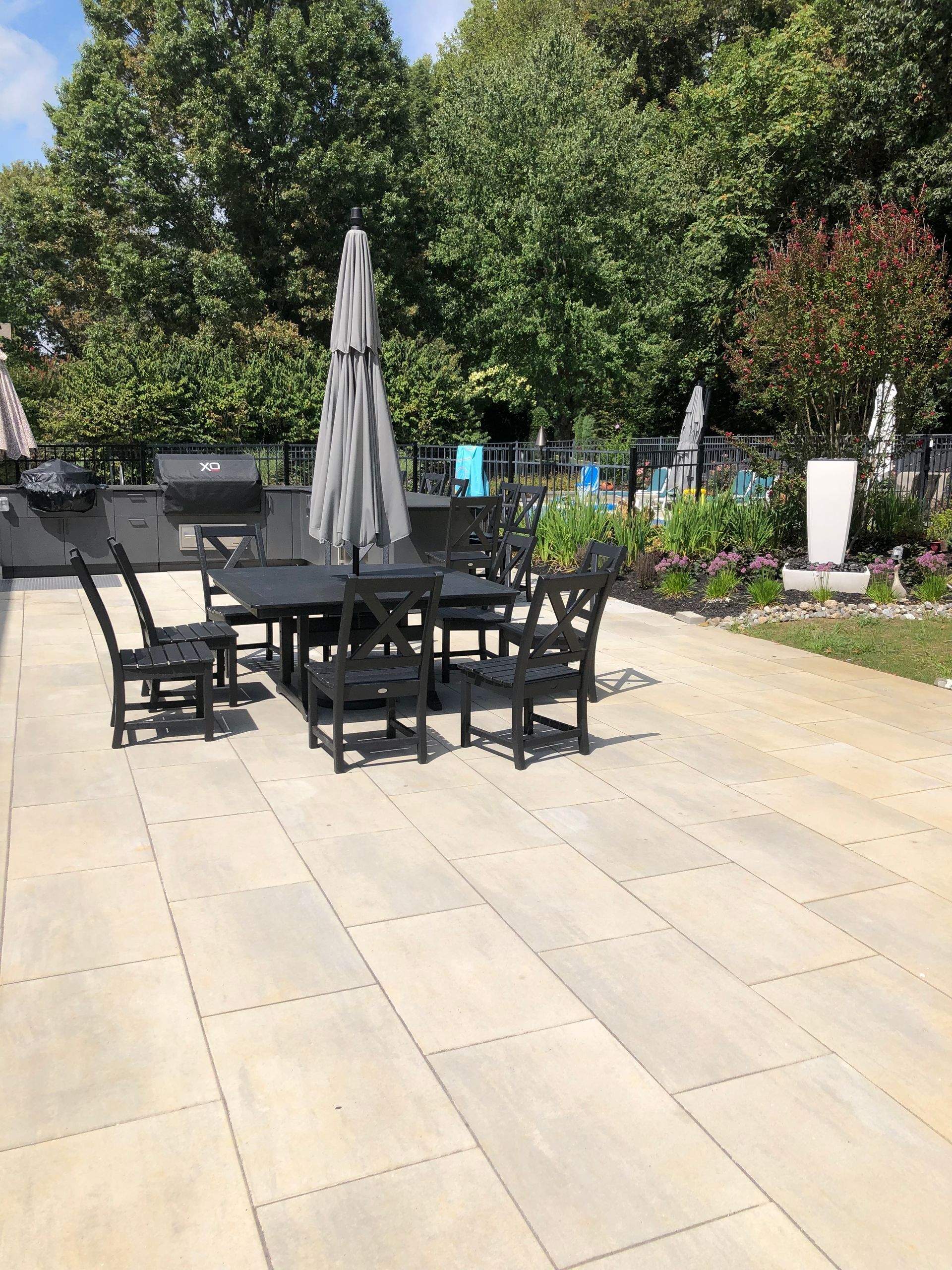 Outdoor patio with black tables and chairs, a gray umbrella, and greenery along the edge