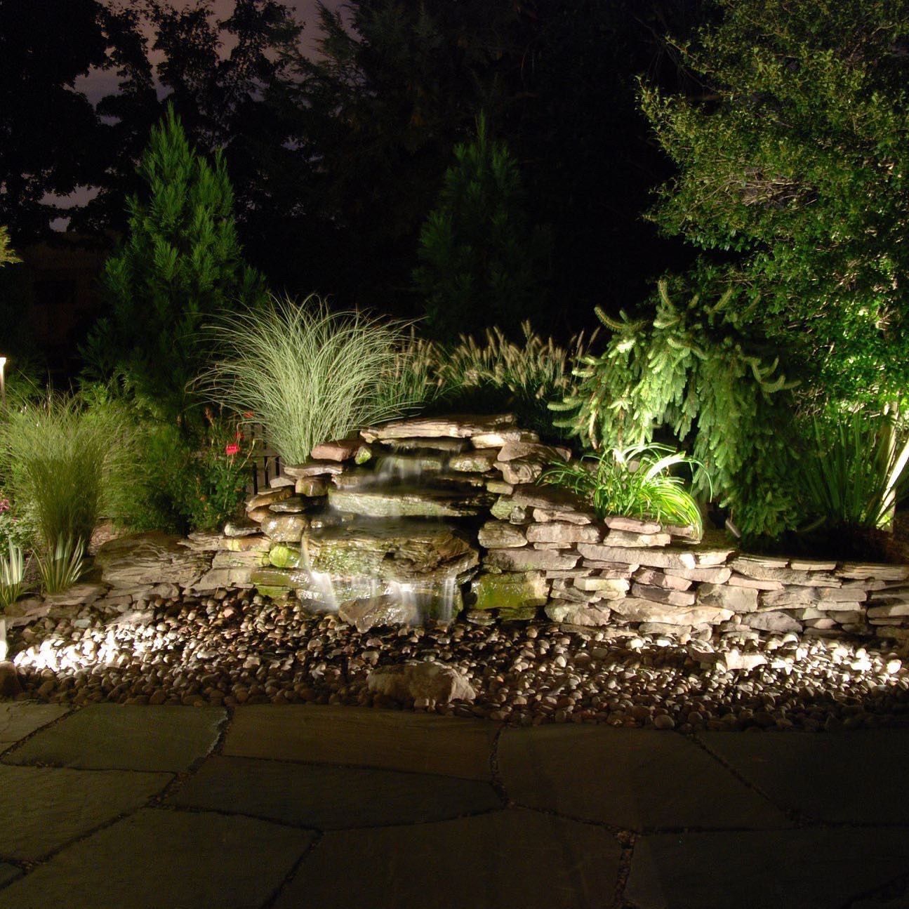 Illuminated backyard stone waterfall with stacked rocks, surrounded by lush greenery at night