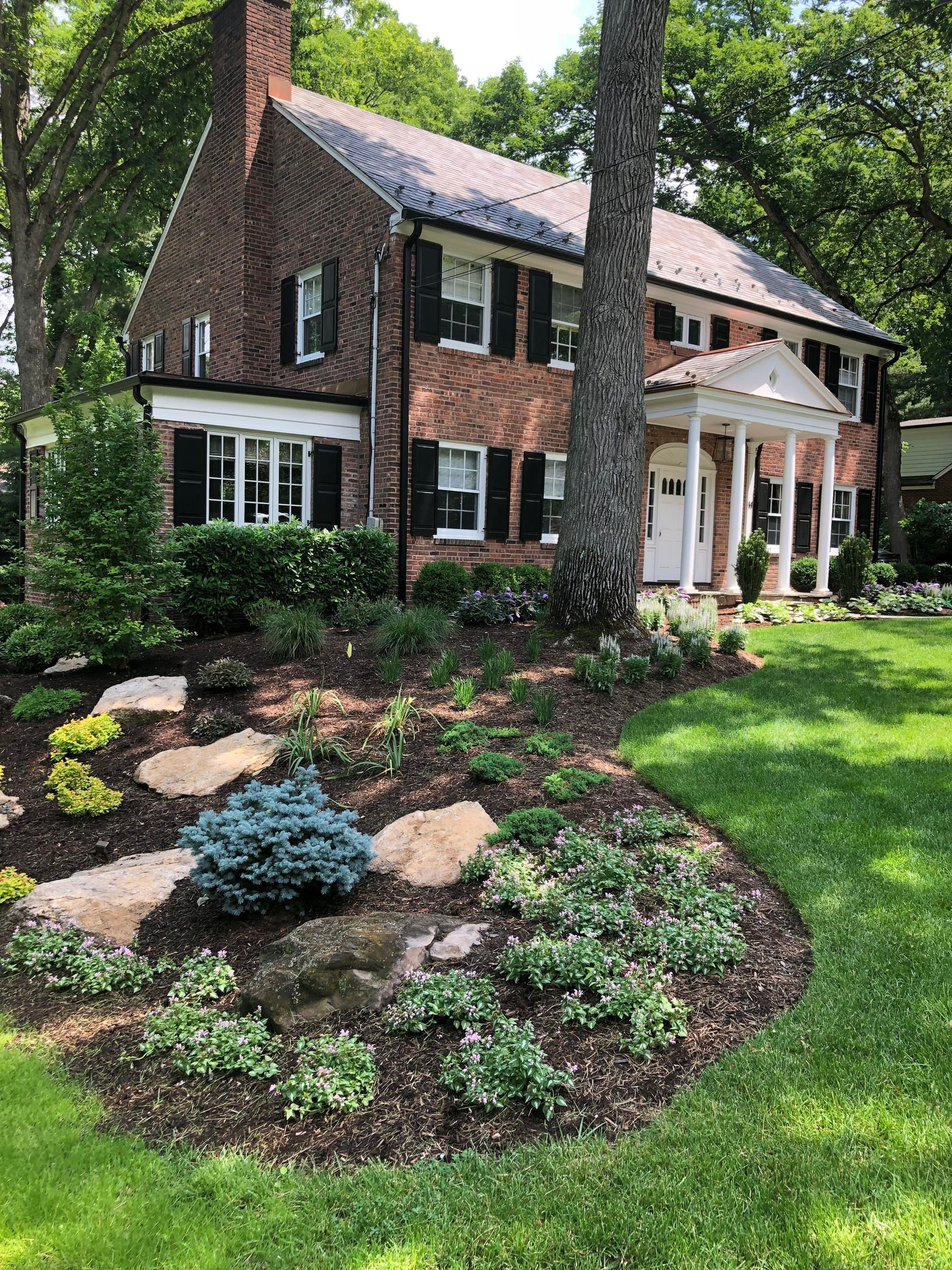 Brick house with white porch, large trees, and landscaped front yard with flowers and lawn