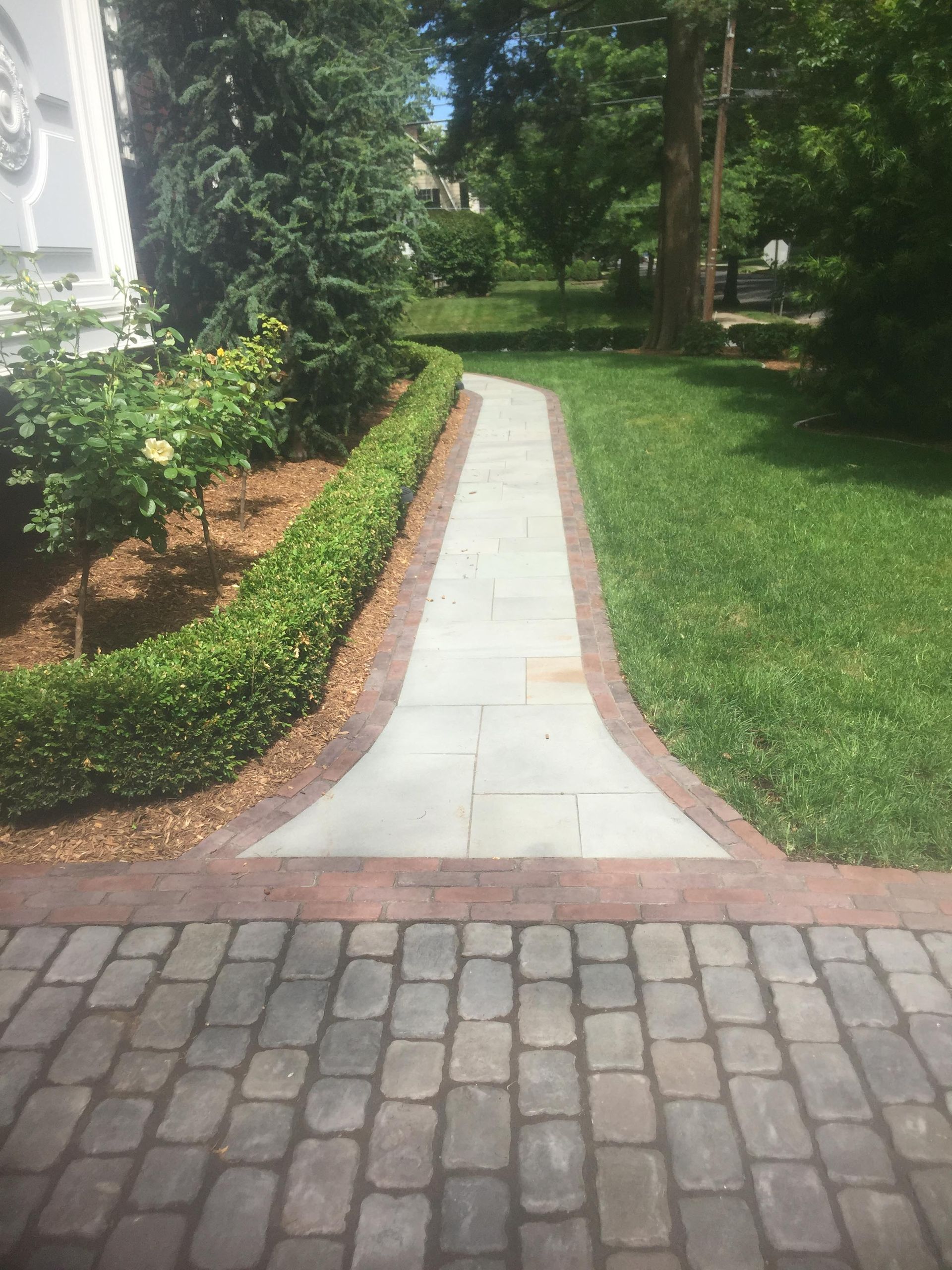 Curved stone walkway beside a house, bordered by red brick and green lawn, leading through a shaded garden.