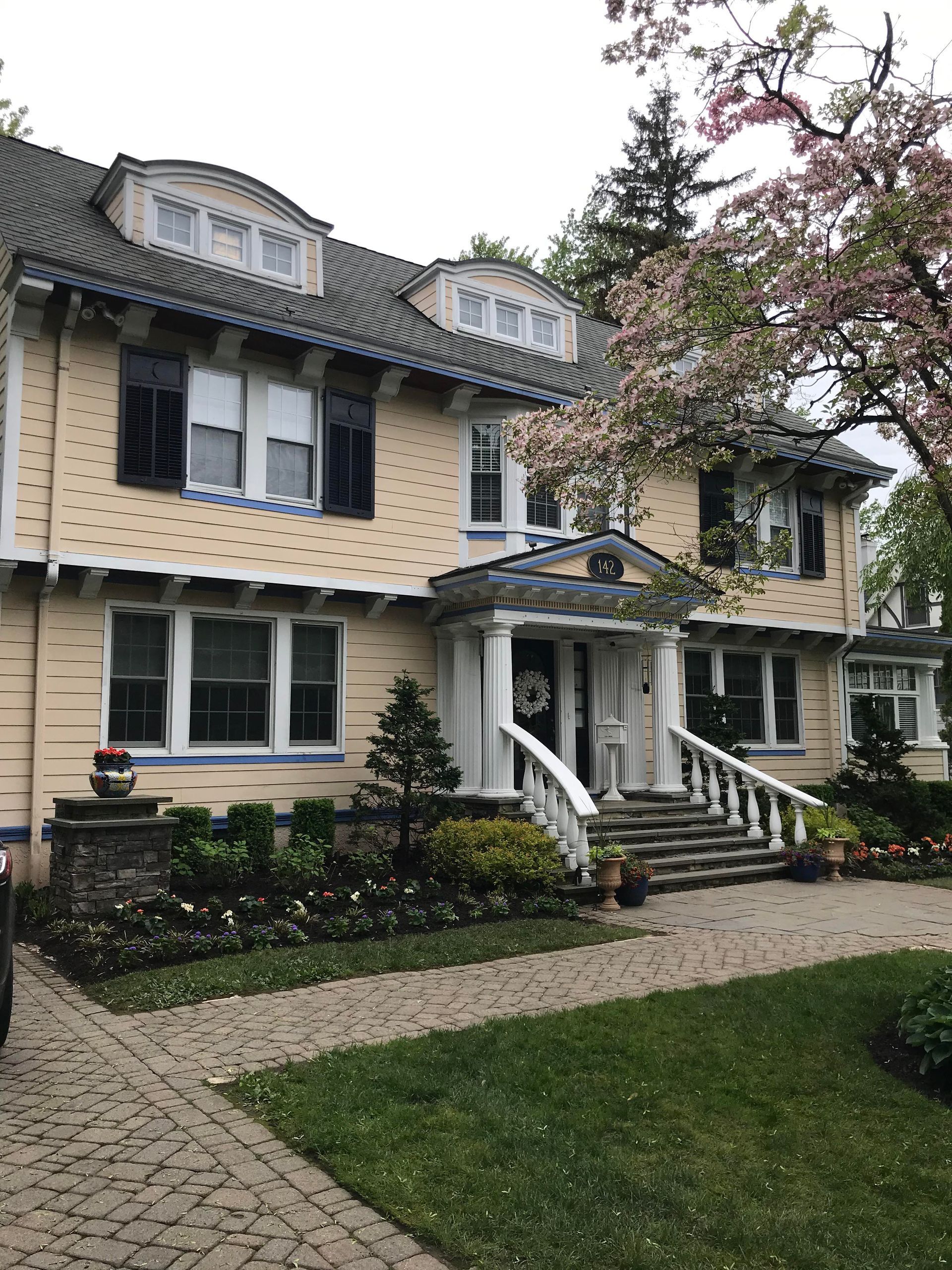 Yellow Victorian house with white trim, dark shutters, front steps, and a garden path beneath blooming trees