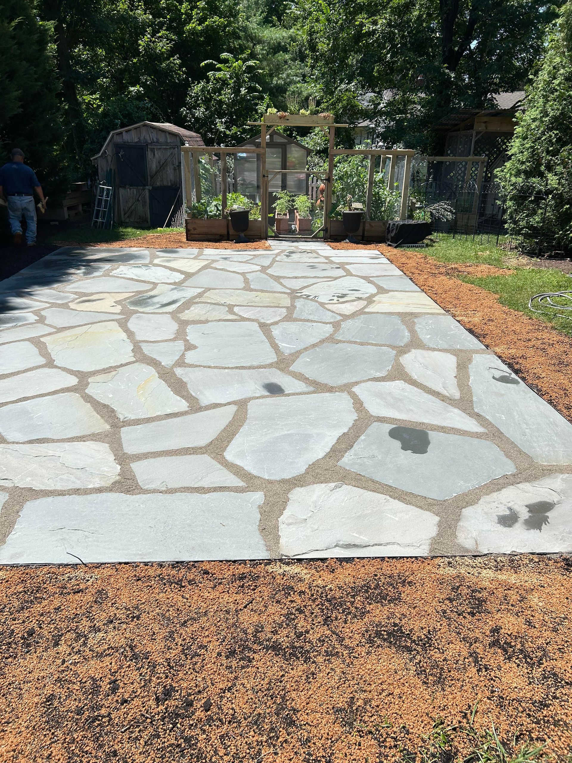 Stone-paved patio with irregular gray flagstones, bordered by mulch and backed by garden fencing and trees.