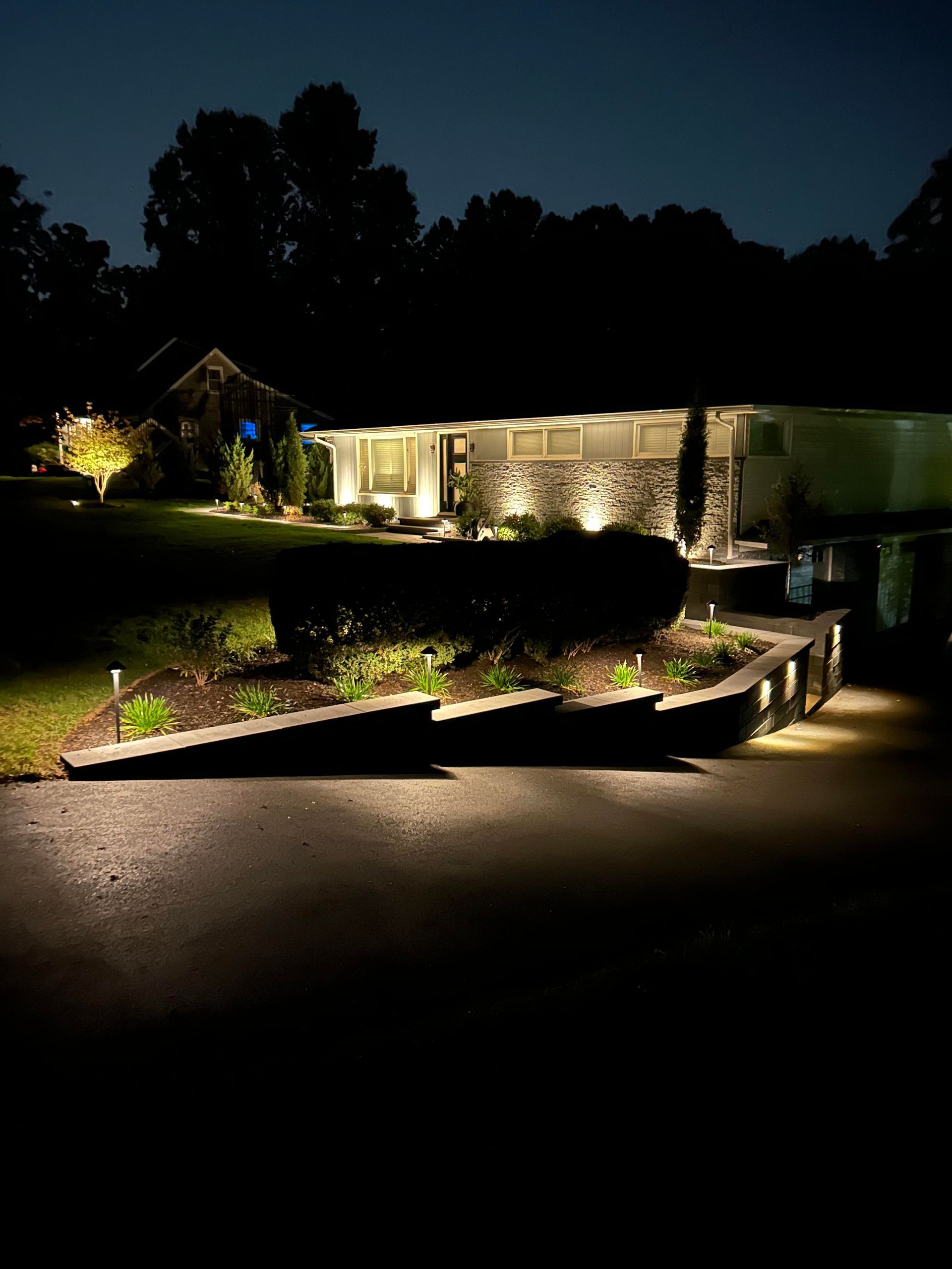 Nighttime suburban house with warm porch lights, landscaped lawn, and driveway lit by ground lights