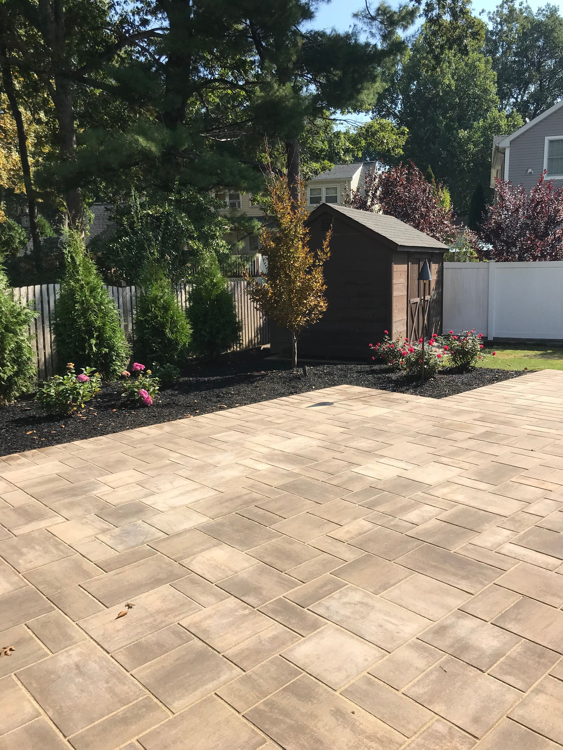 Sunny backyard patio with tan pavers, garden beds, shrubs, and a small shed beside a white fence.