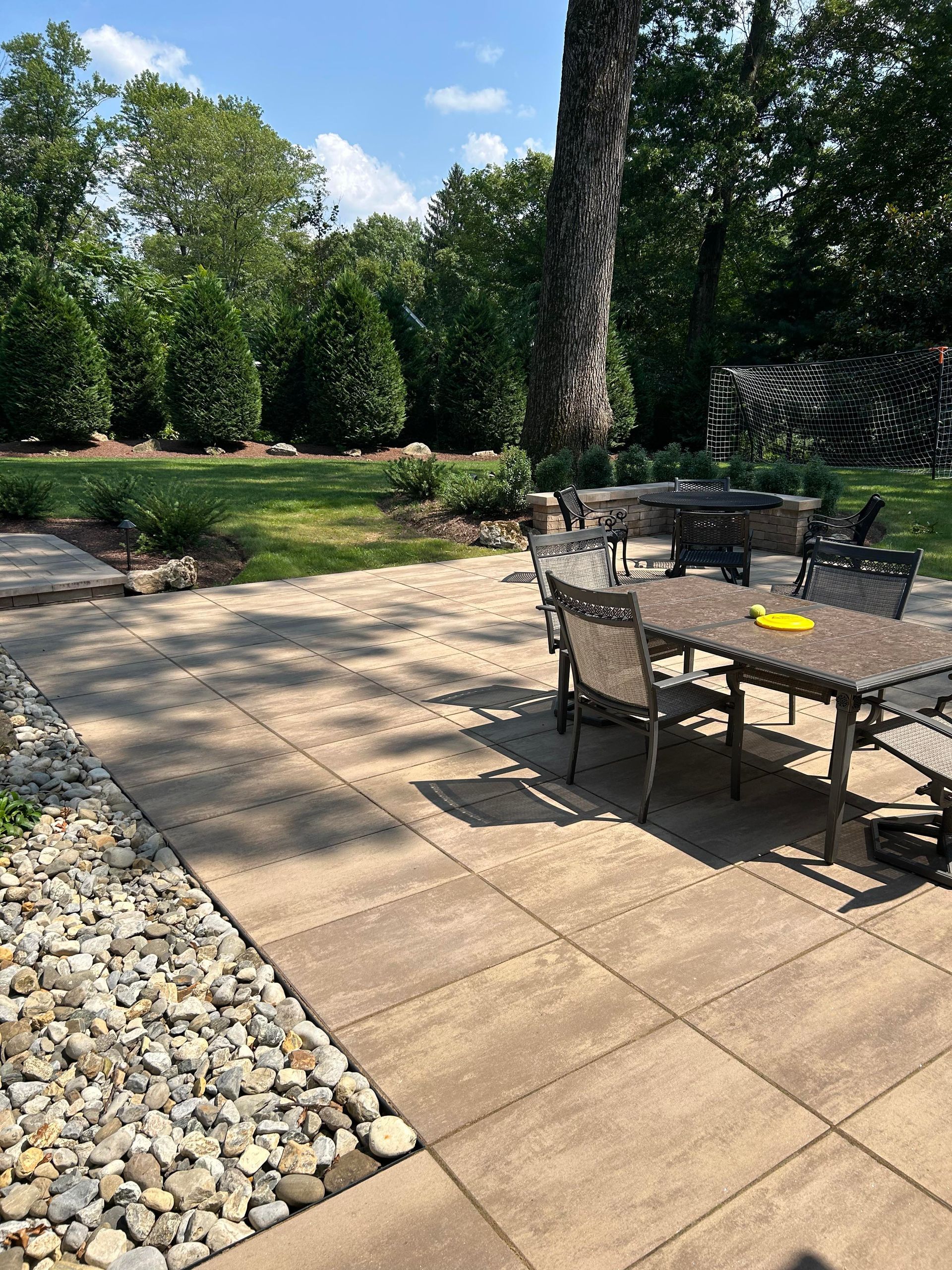 Patio with wicker table and chairs beside a landscaped garden, stone path, and trees under a blue sky