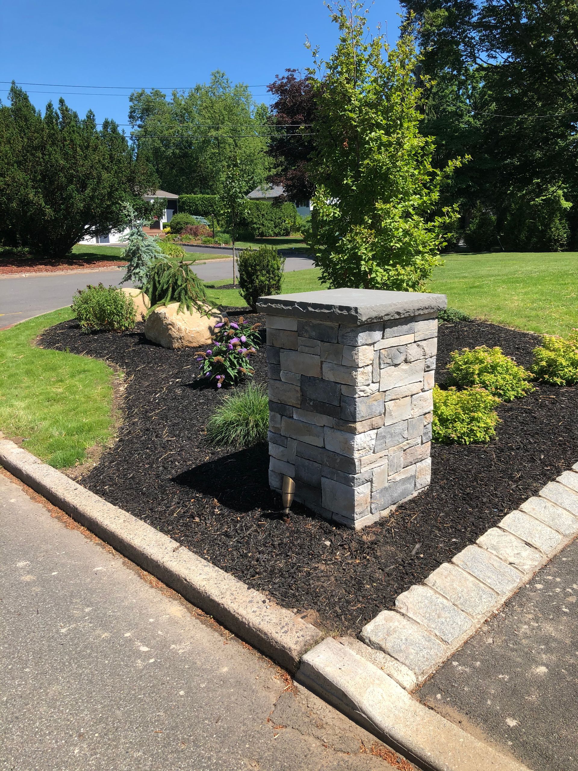 Landscaped median with shrubs, a small tree, and a stone pillar beside a paved road