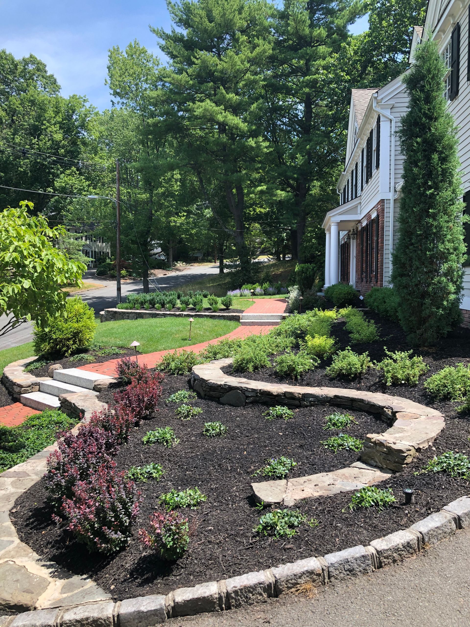 Landscaped front yard with curved stone edging, shrubs, and a white house by a tree-lined driveway