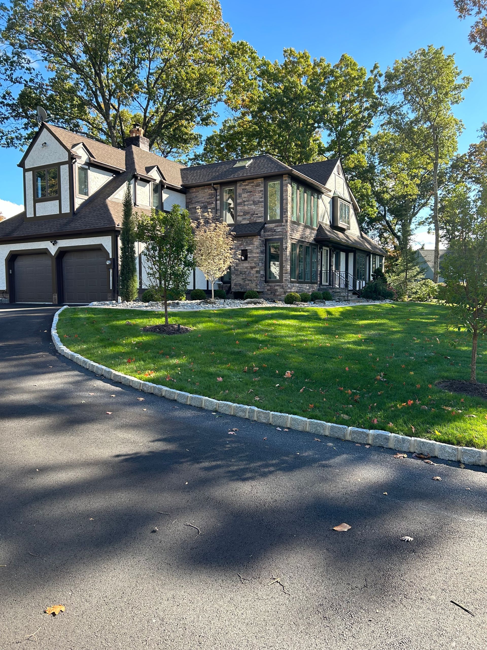 Stone suburban house with two garages, curved driveway, and green lawn under sunny trees