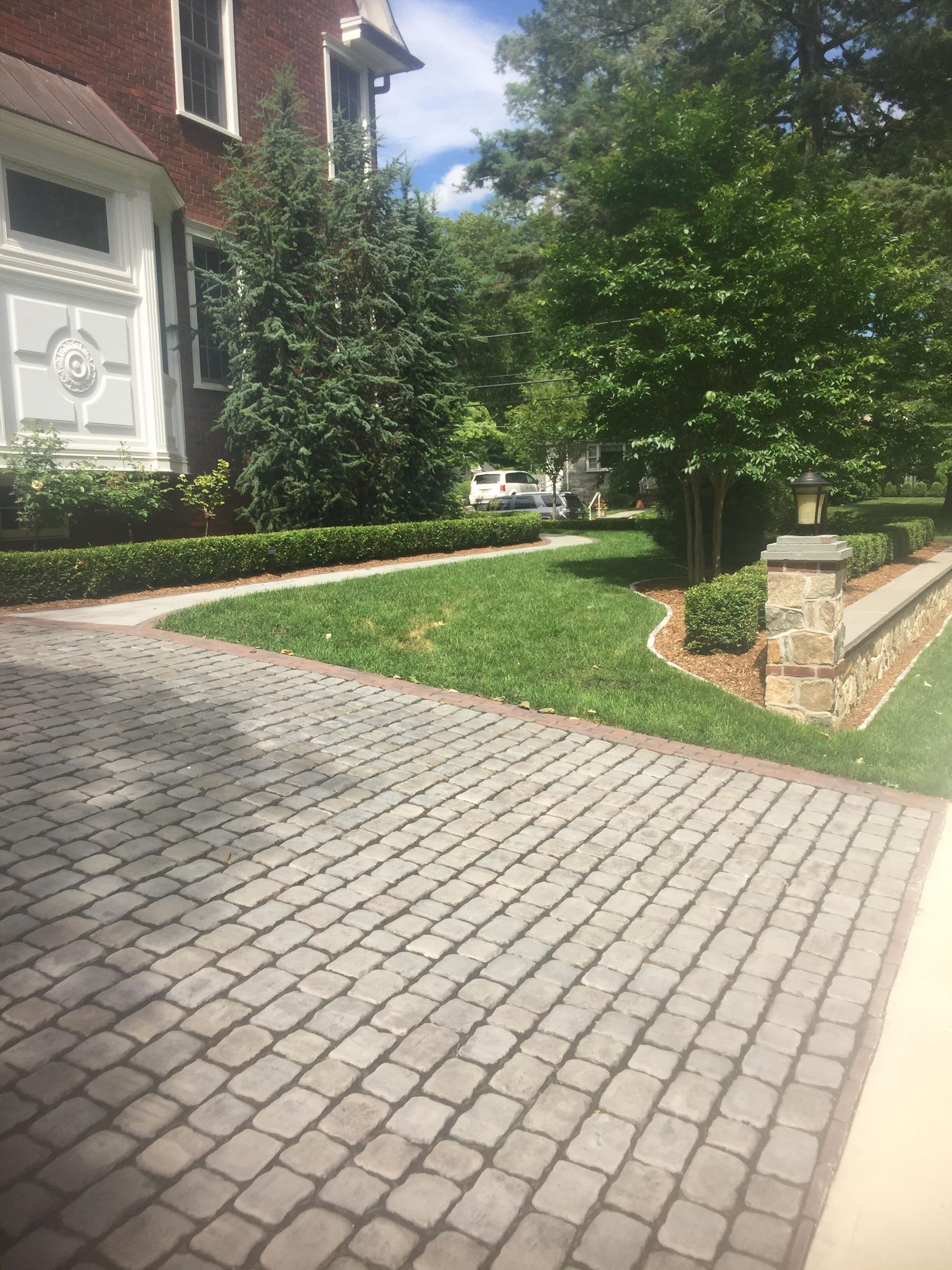 Brick driveway beside a manicured lawn and shrubs next to a house on a sunny day