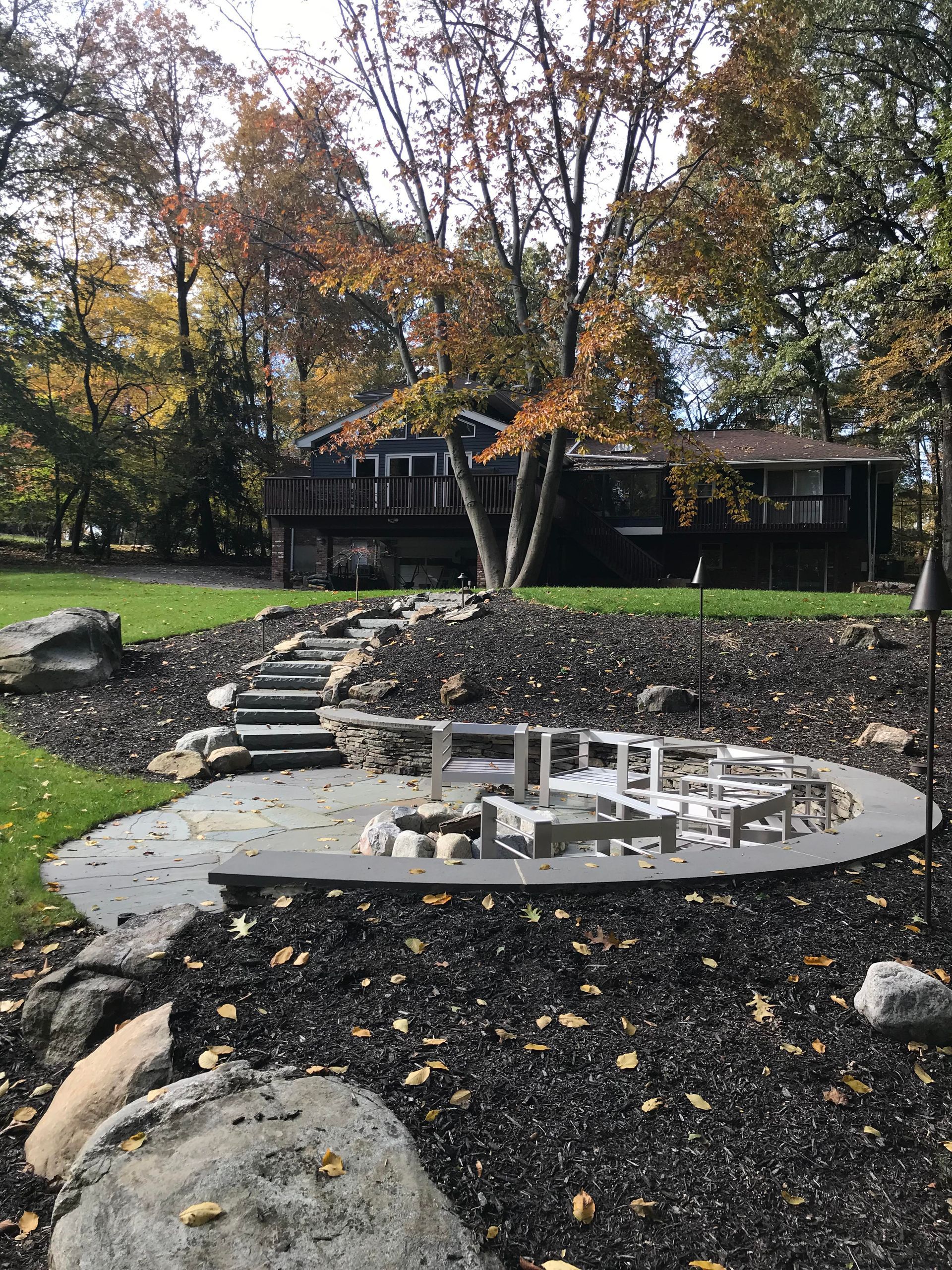 Curved stone fire pit in a landscaped yard with steps, trees, and a dark house in the background