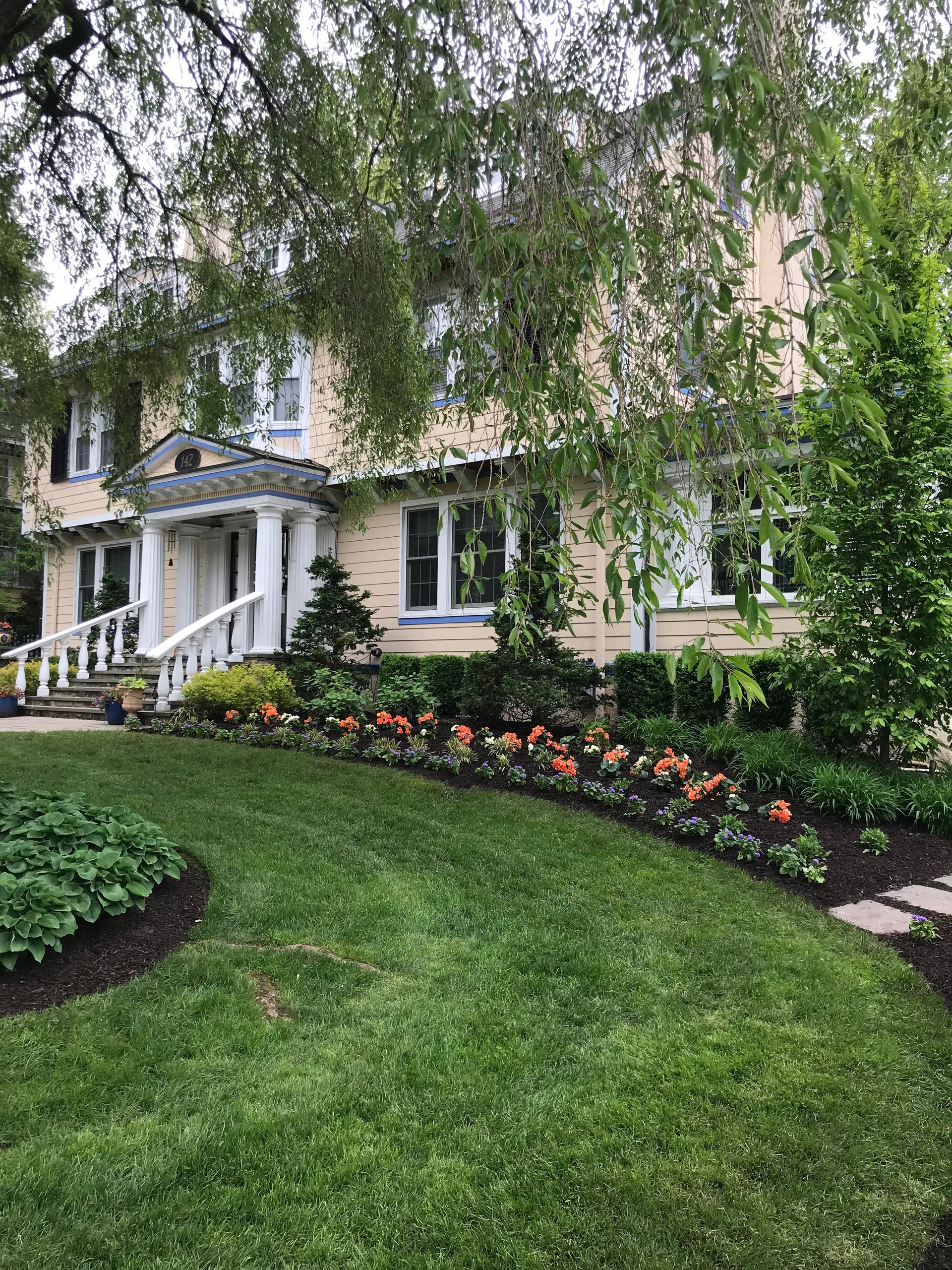 Landscaped cream-colored house with white porch, lush green lawn, and flower beds under hanging tree branches