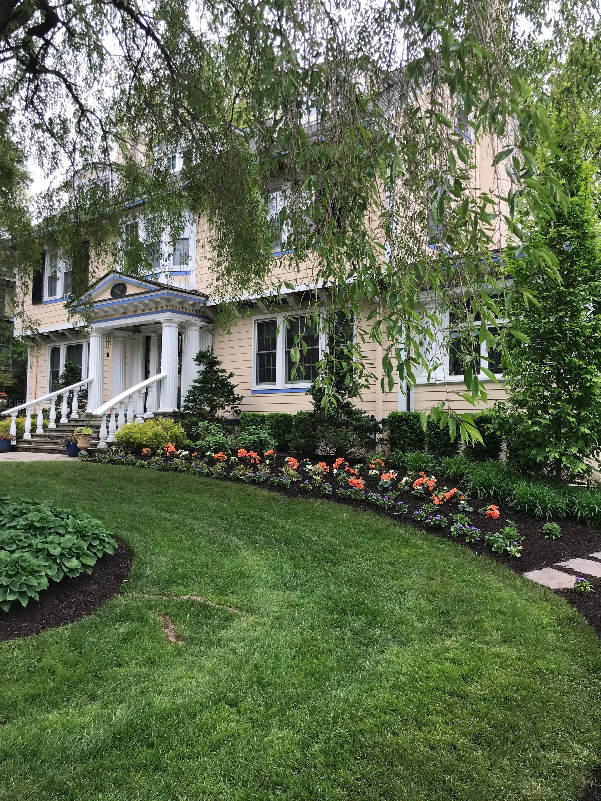 Suburban house with white columns, lush green lawn, flowering beds, and drooping tree branches