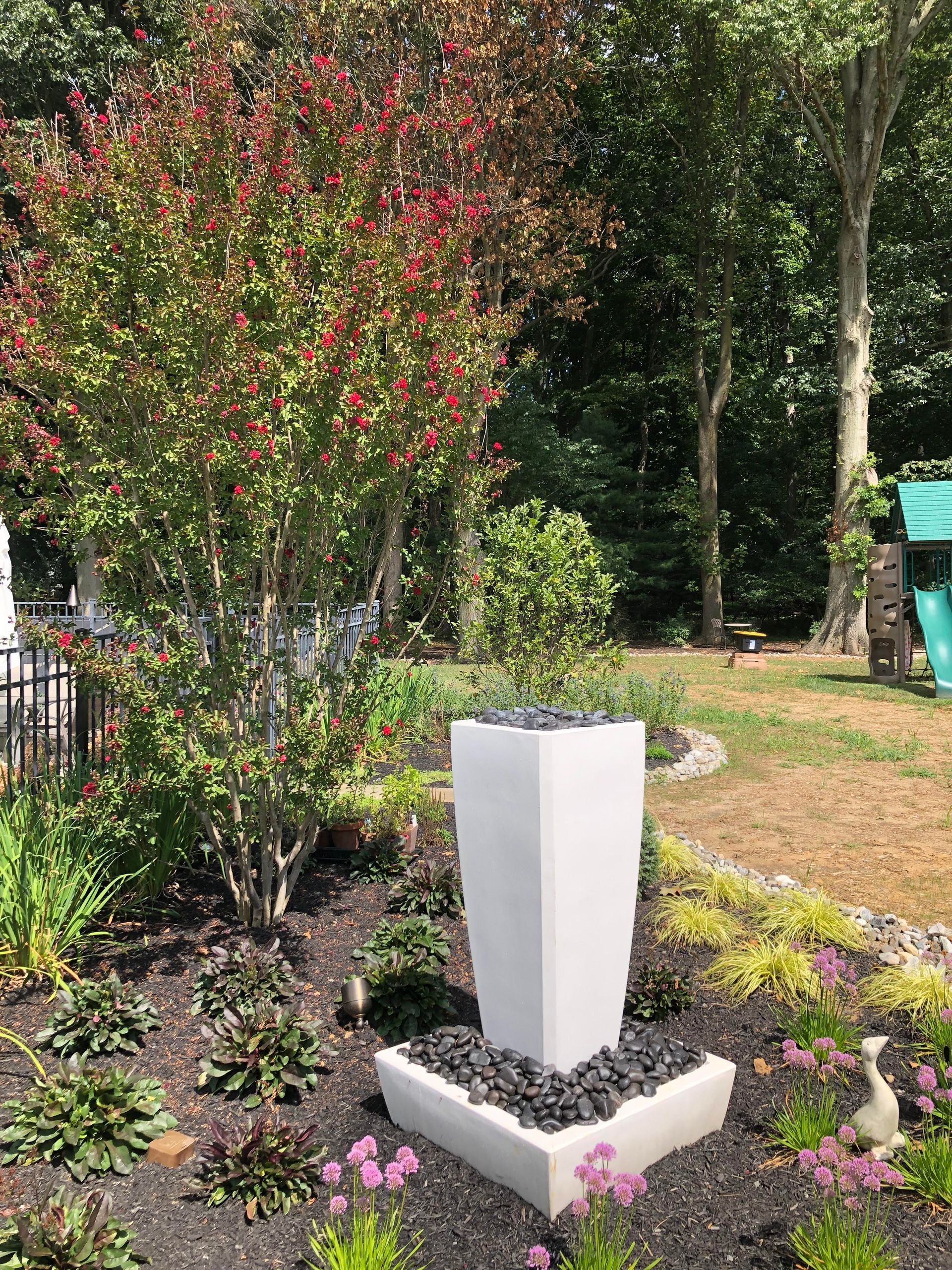 White memorial pedestal in a garden bed with pink flowers and trees in the background.