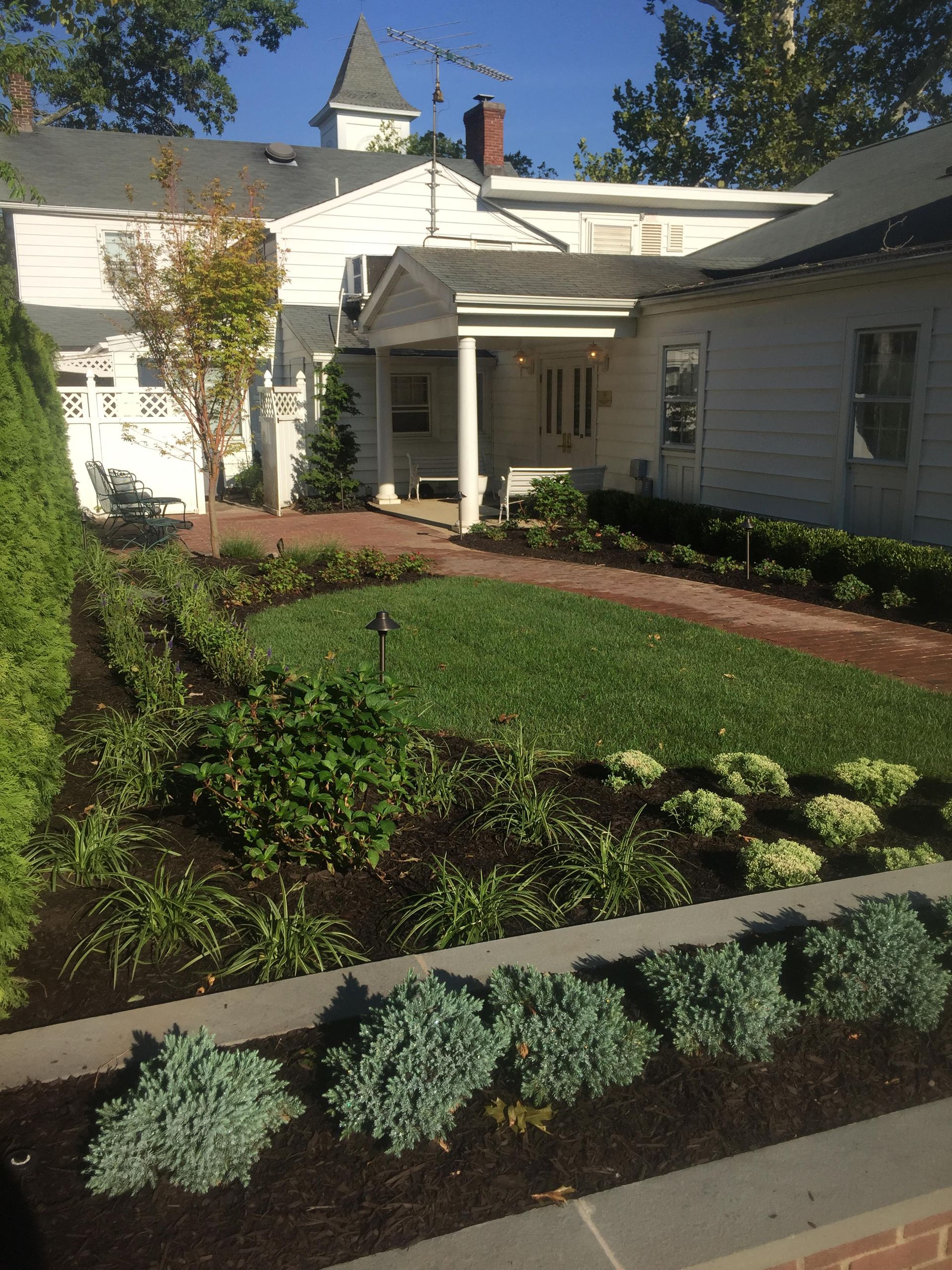 White house with front porch and landscaped garden beds under a clear blue sky.