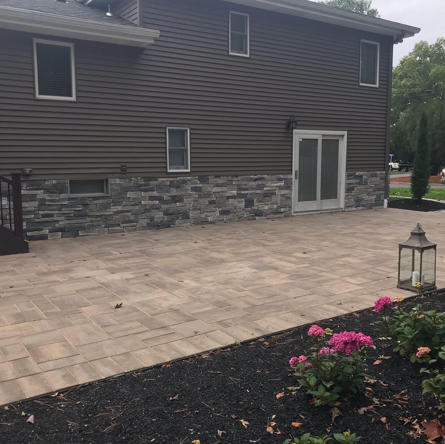 Stone patio beside a dark-sided house with sliding doors, lantern, and pink flowers in the foreground.