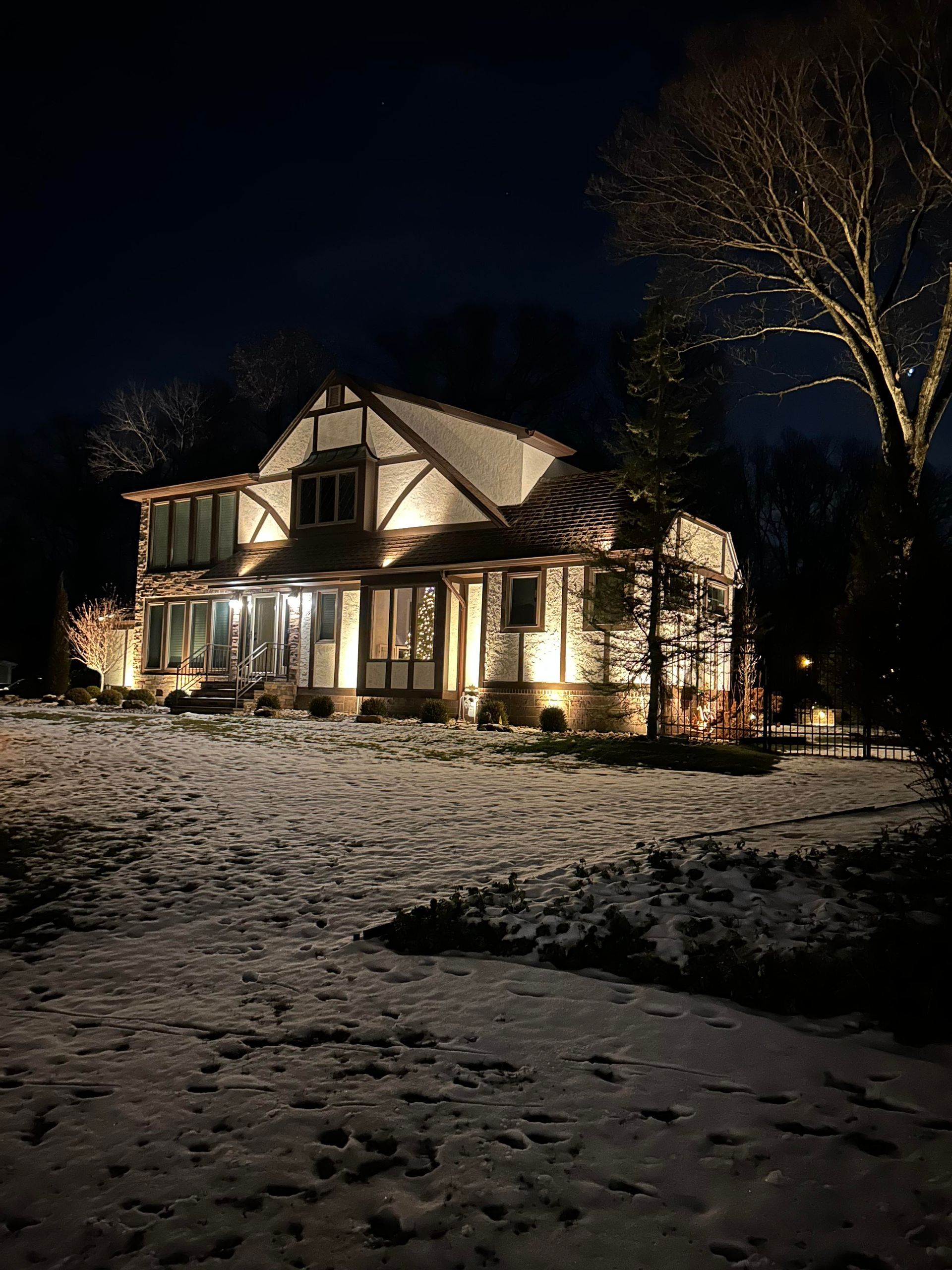 Snow-covered house lit warmly at night under a dark sky, with bare trees nearby