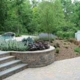 Landscape with curved stone planter, stairs, and green shrubs beside a paved walkway