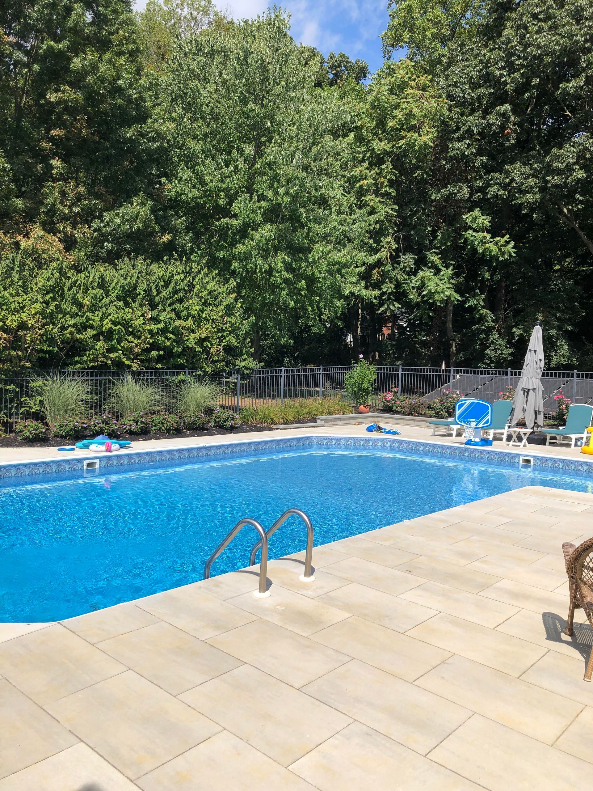 Outdoor swimming pool with blue water, beige deck, lounge chairs, and dense green trees around it