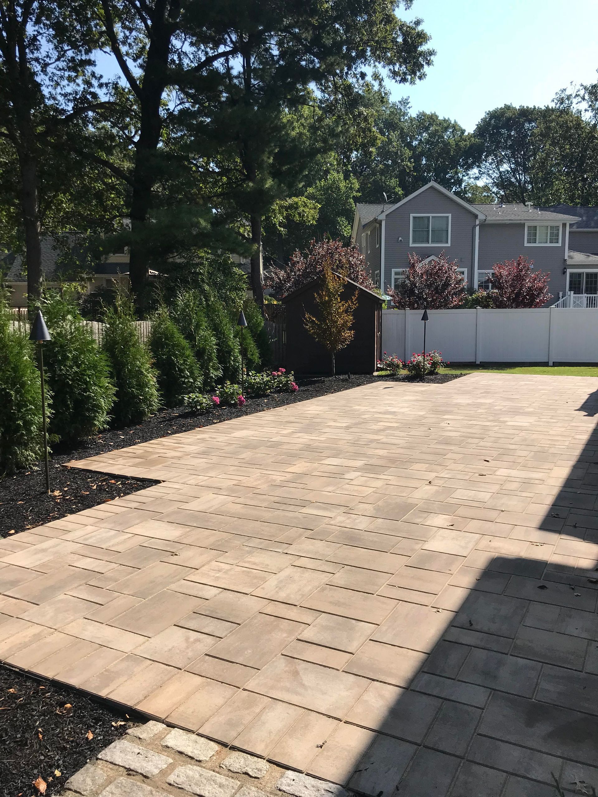 Brick patio in a sunny backyard with trees, shrubs, and a house in the background.