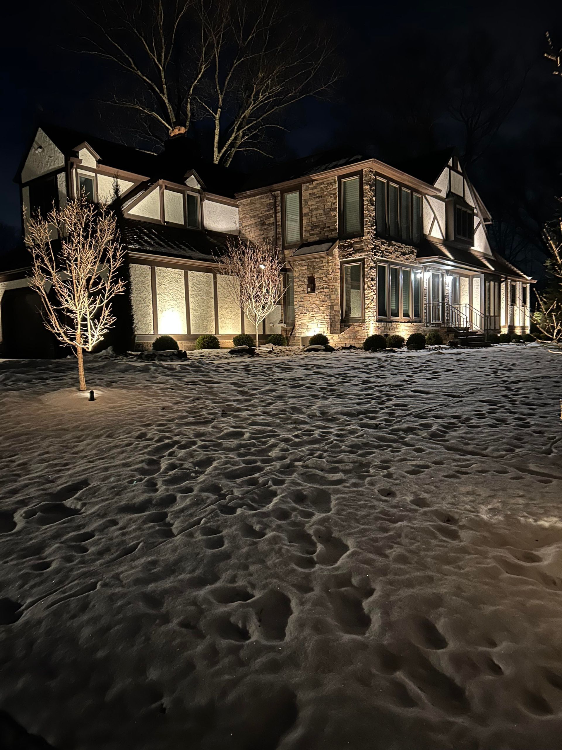 Snowy front yard at night with a lit two-story house and glowing windows