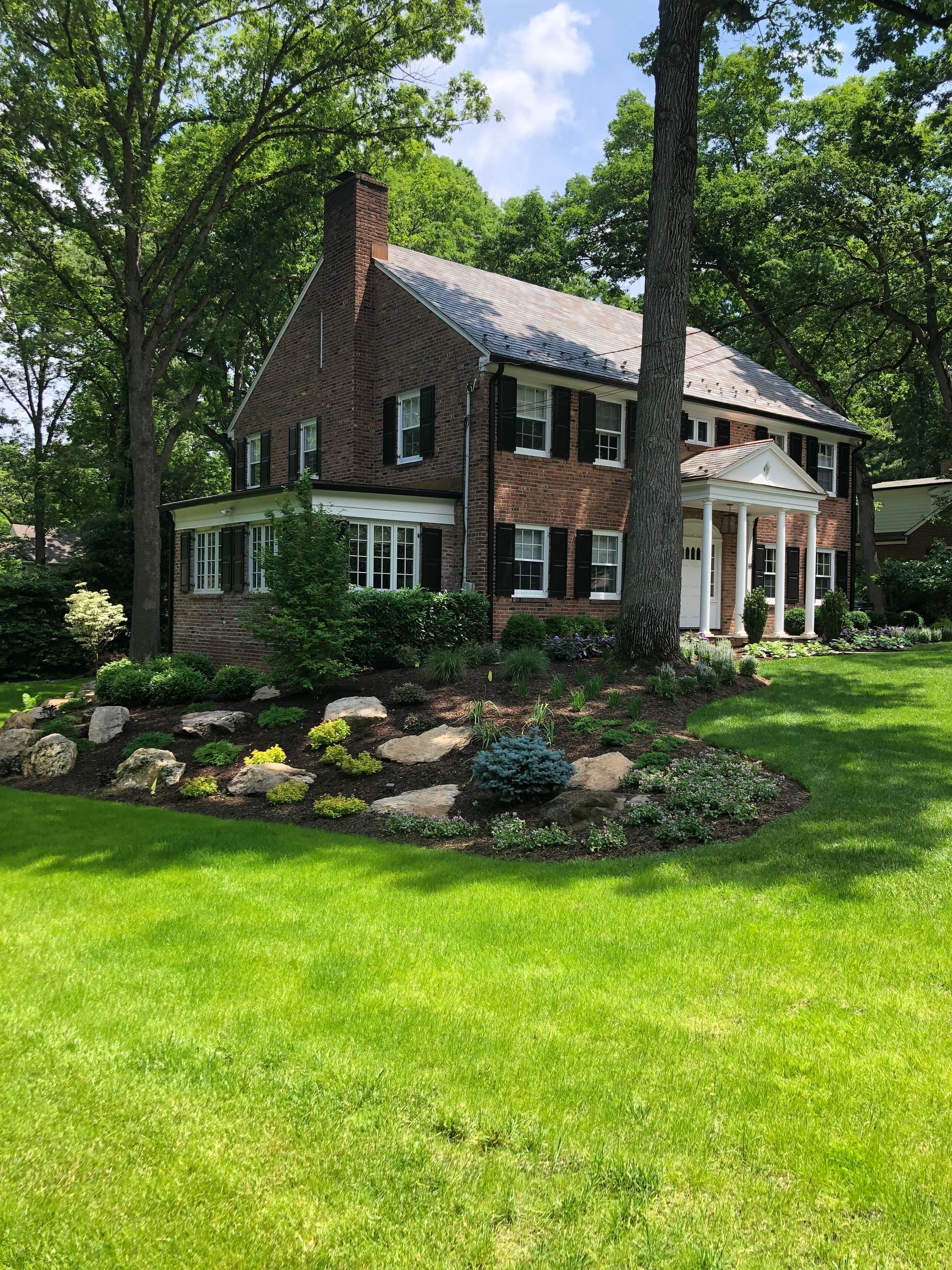 Brick house with white columns and chimney, set in a lush green lawn and landscaped garden under trees.