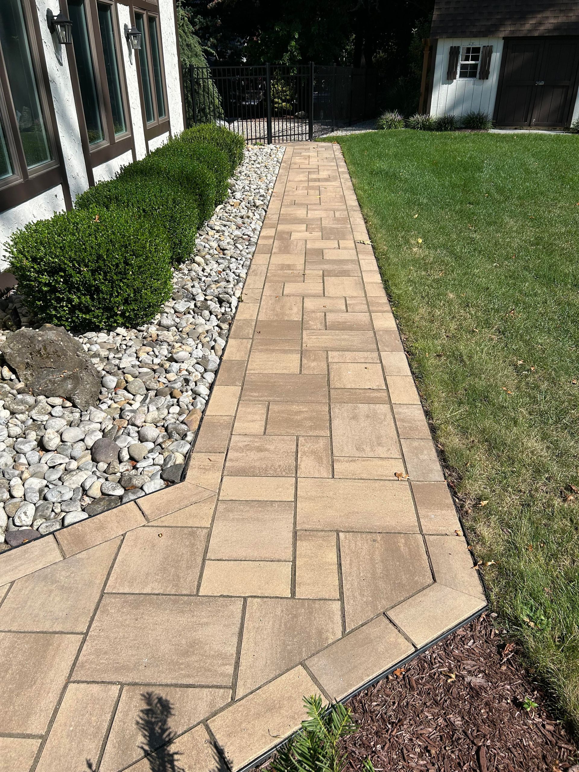 Tan brick paver walkway through a landscaped yard, bordered by rocks, shrubs, and green grass.