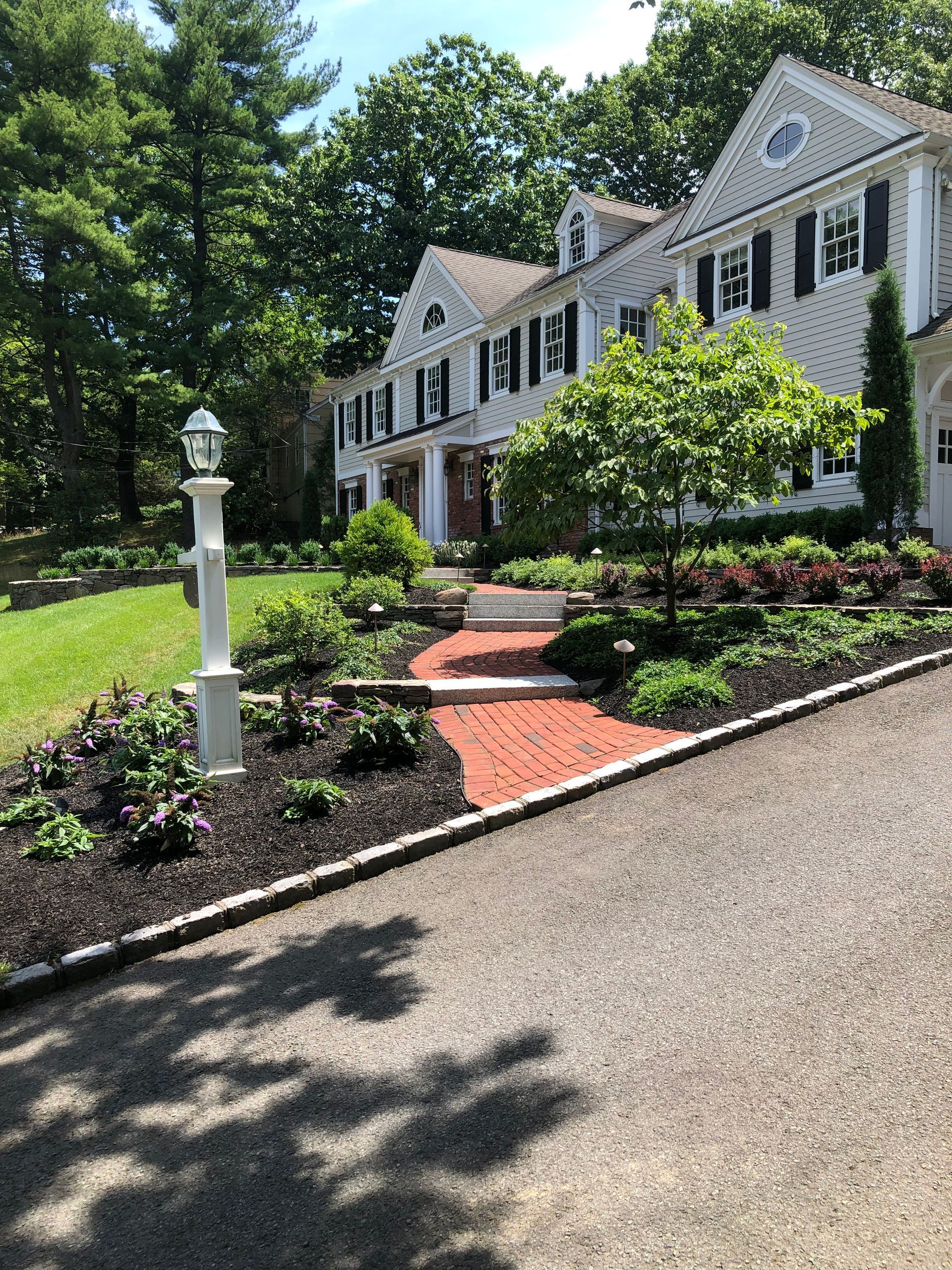 White colonial-style house with black shutters, curved brick walkway, and landscaped front yard under trees