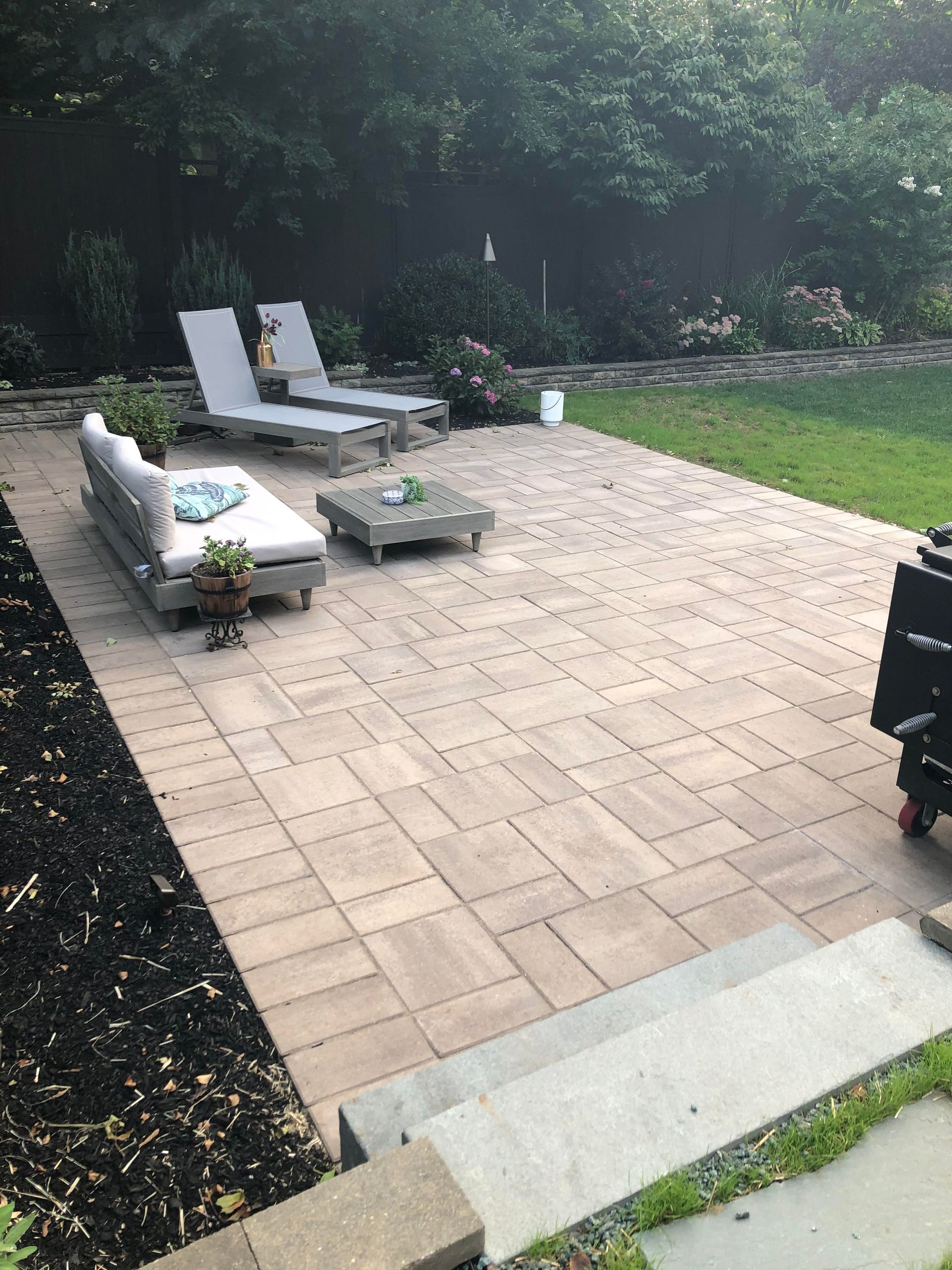 Patio with outdoor seating, white lounge chairs, and a fire pit beside a grassy yard and trees.