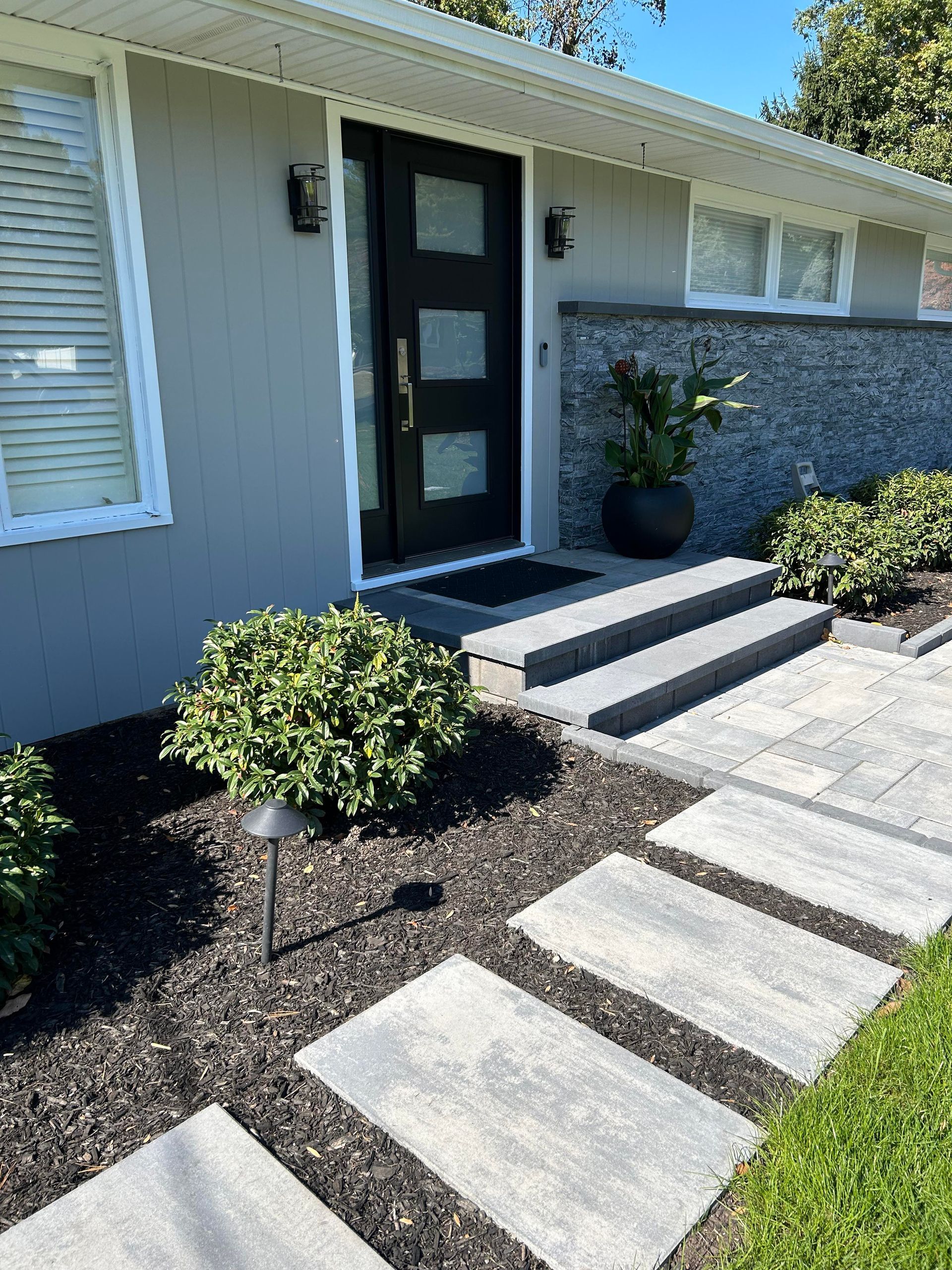 Front entry walkway with concrete steps, black door, and gray siding beside a landscaped porch.