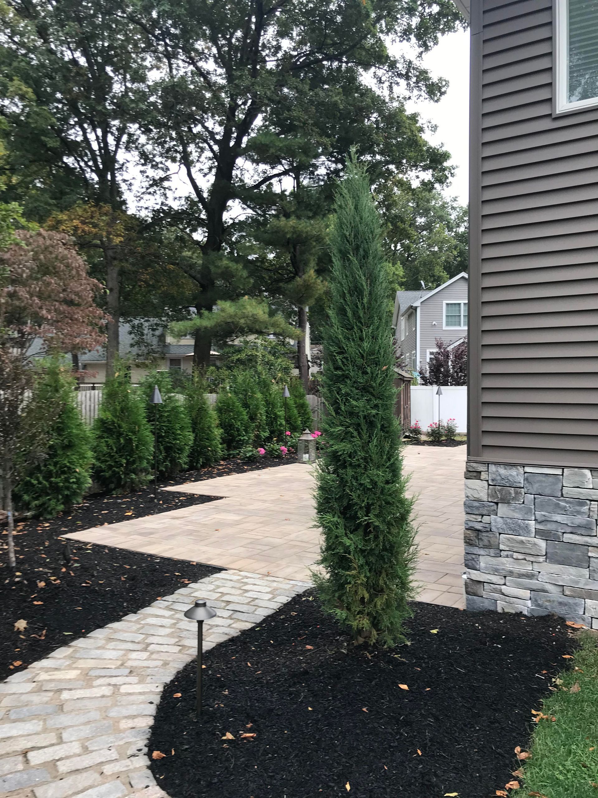 Curved stone walkway through black mulch and tall shrubs beside a gray house