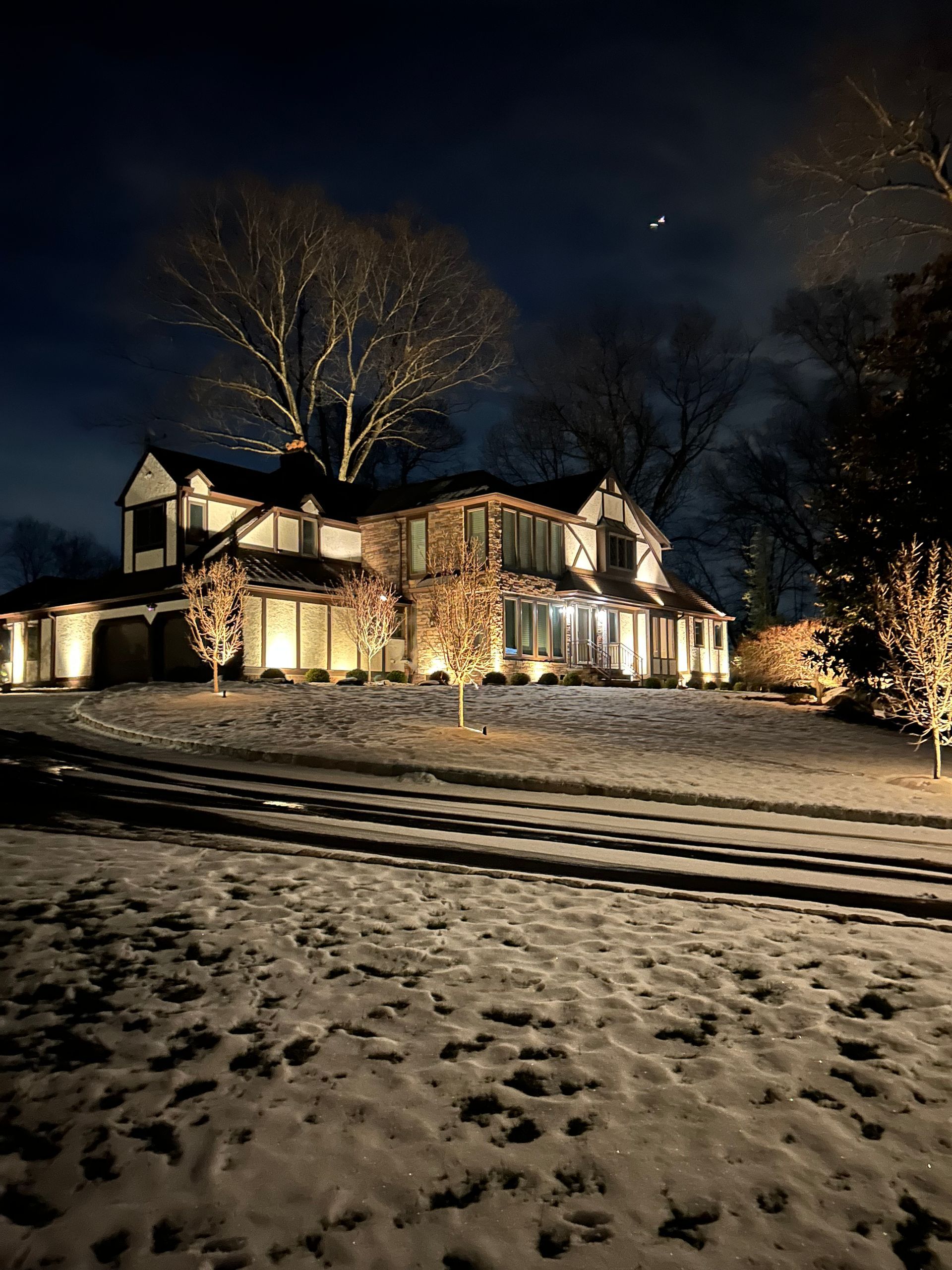 Snowy yard with a warmly lit house at night and bare trees under a dark sky