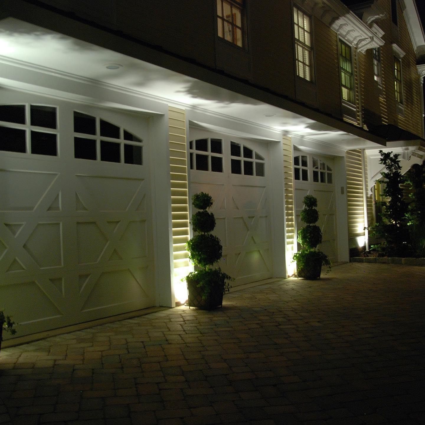 Nighttime view of a brick driveway with lit white garage doors and windows on a house facade