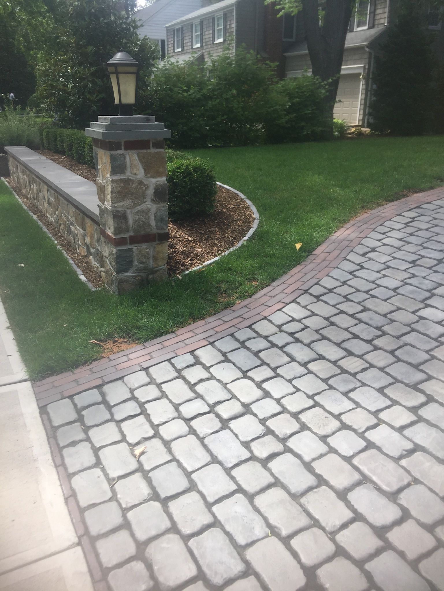 Brick-paved driveway beside a stone garden wall, landscaped lawn, and house exterior